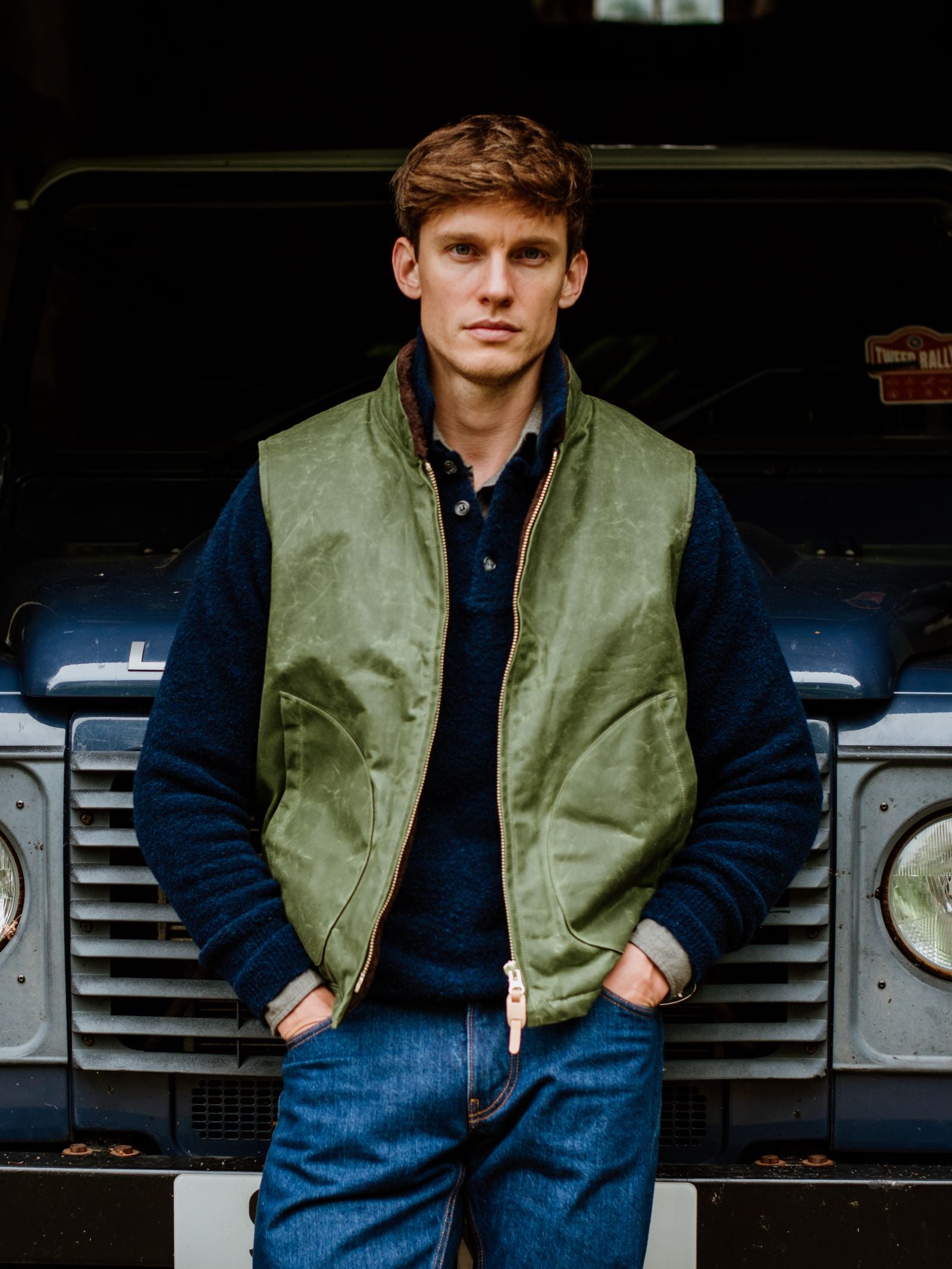 A young man with short brown hair wears a Campbell's of Beauly Wax Nehru Gilet over a blue sweater, standing with hands in his pockets before a dark blue vehicle, looking directly at the camera.