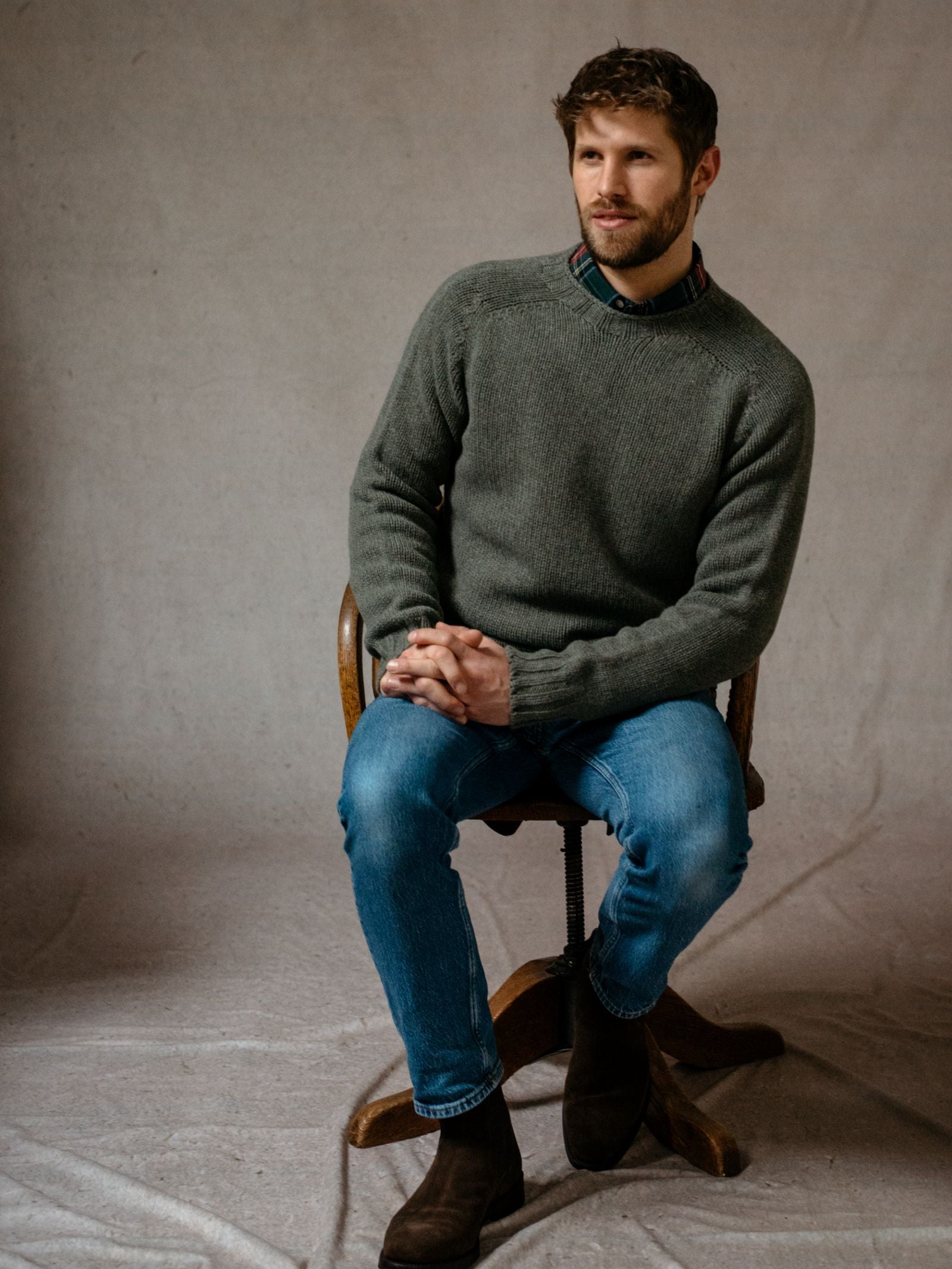 A man with short brown hair and a beard sits on a wooden chair, hands clasped, wearing the Campbells of Beauly Geelong Lambswool Chunky Crew Jumper in green over a plaid shirt, blue jeans, and brown boots against a neutral backdrop.