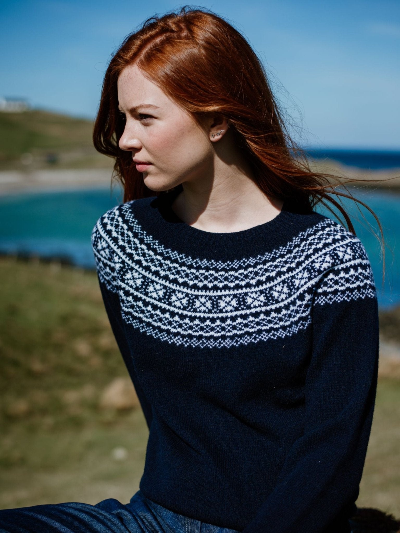 A woman with long red hair wears the Two-Colour Fairisle Yoke Jumper by Campbells of Beauly, a navy Geelong lambswool sweater with a traditional yoke, as she sits outdoors by green grass, water, and a blue sky.
