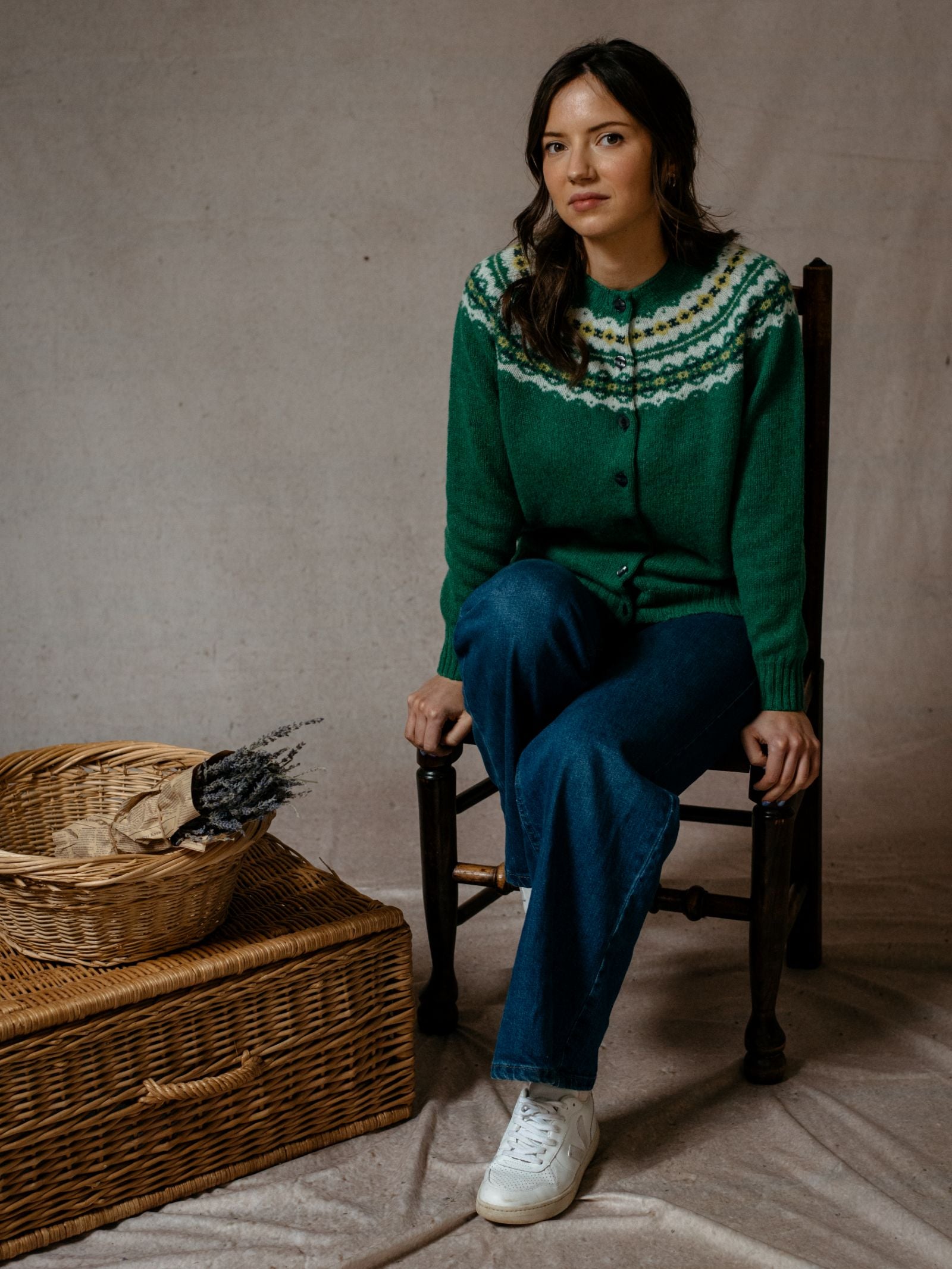 A woman with long dark hair, wearing the Campbell’s of Beauly Shetland Fairisle Cardigan, blue jeans, and white sneakers, sits on a wooden chair next to two woven baskets against a neutral fabric backdrop.