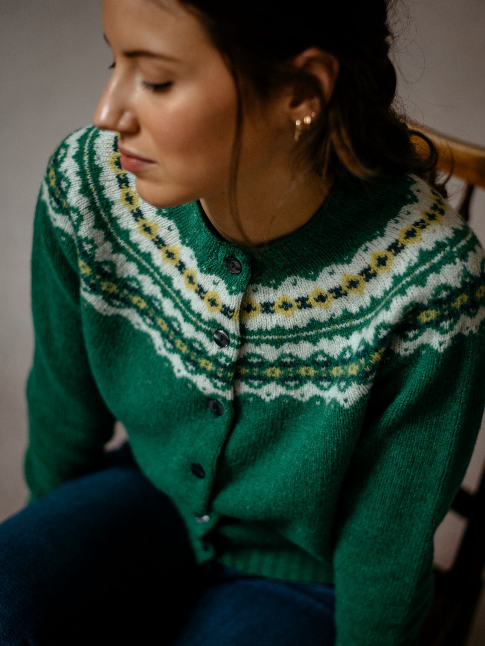 A woman with brown hair, gold hoop earrings, and a green Shetland Fairisle Cardigan by Campbells of Beauly featuring white and yellow details sits on a chair, looking down calmly.