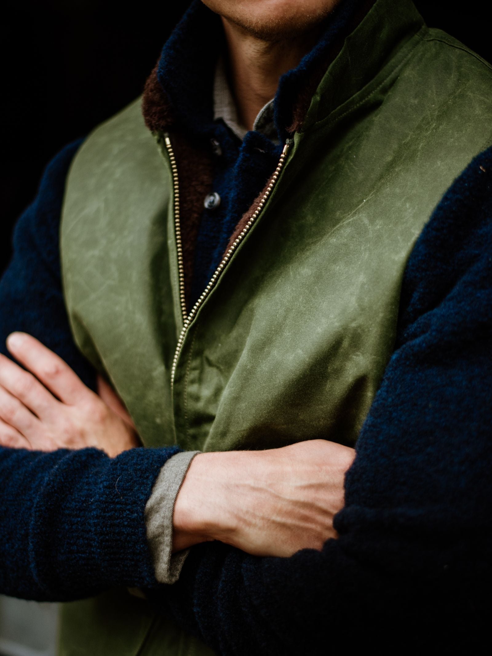 A torso shot shows a person with arms crossed, wearing Campbell's of Beauly's Wax Nehru Gilet in green over a dark blue fleece jacket with Nehru collar and a gray shirt.