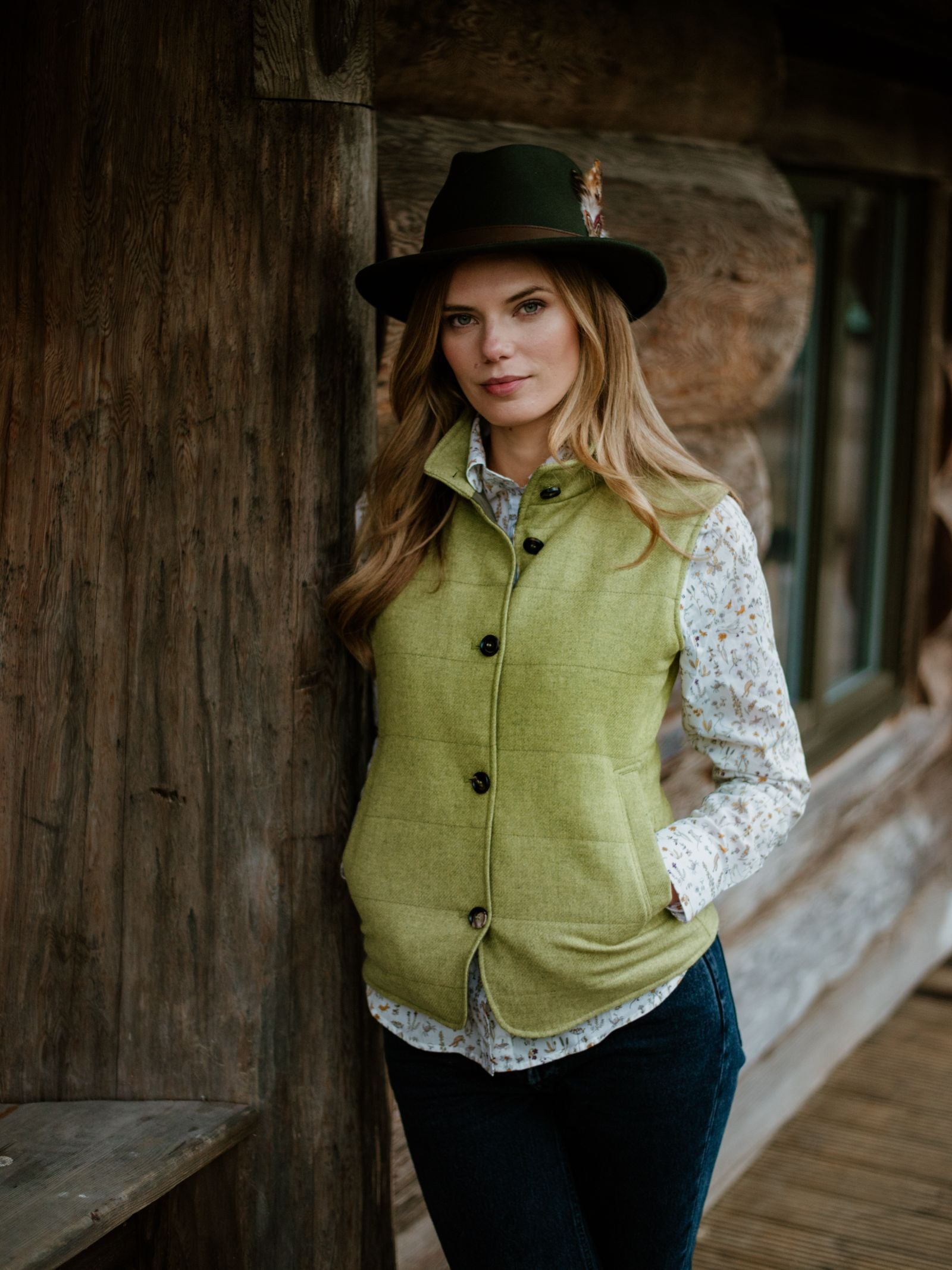 A woman wears the Campbells of Beauly Quilted Buttoned Gilet over a patterned shirt, dark jeans, and a feathered hat, leaning against a wooden cabin wall—a versatile layering piece inspired by British tweed style.