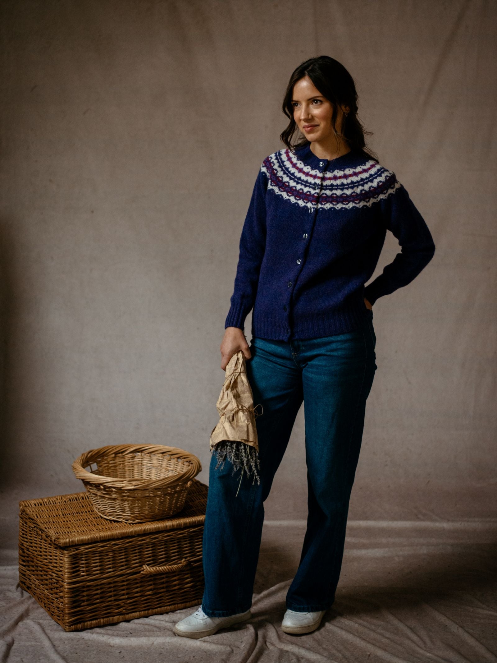 A woman wears Campbells of Beauly Shetland Fairisle Cardigan and jeans, holding a cloth beside two wicker baskets indoors, with a neutral brown backdrop.