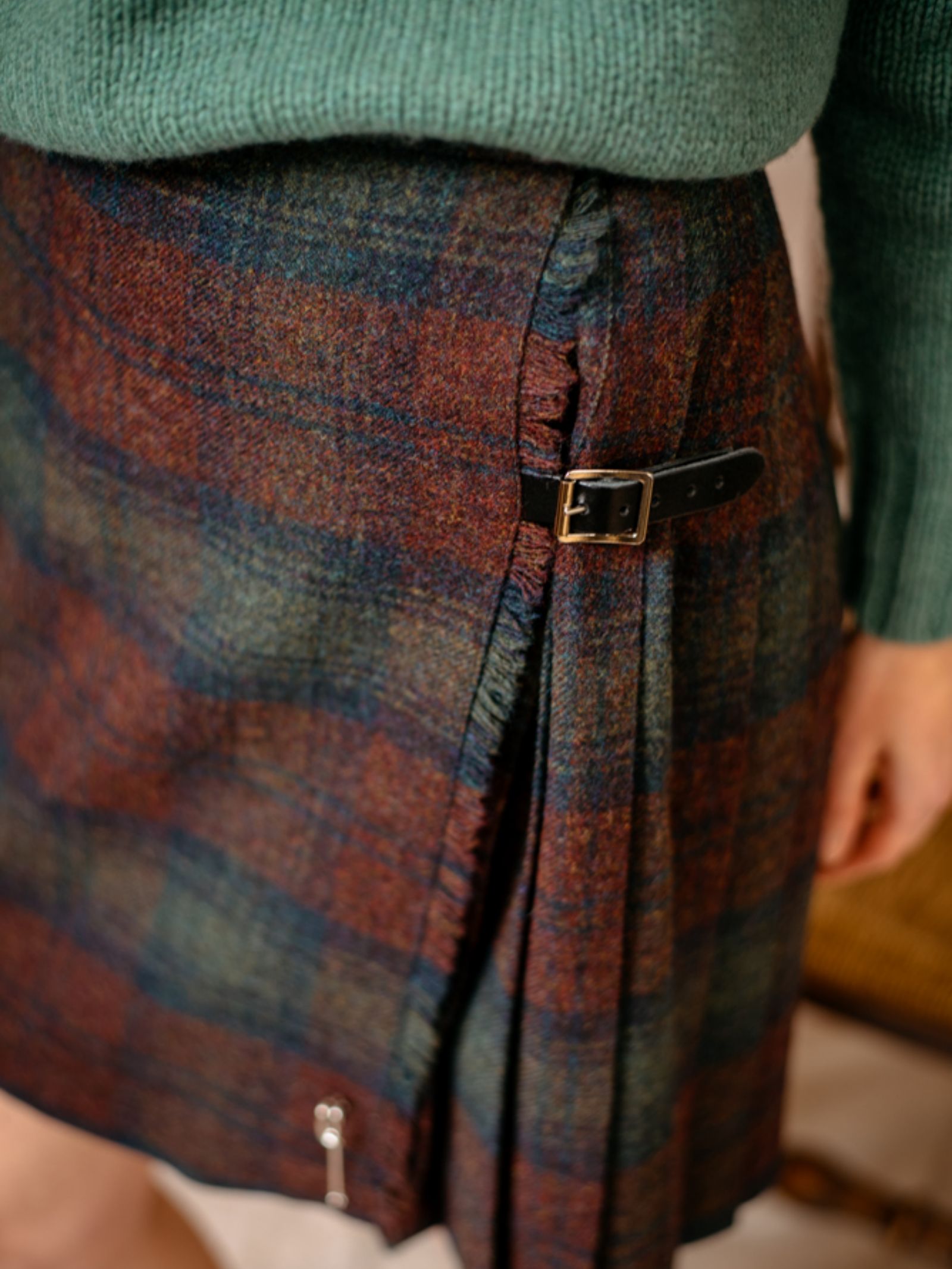 A close-up shows the lower torso and arm of a person wearing a green sweater and Campbells of Beauly Tweed Kilt—a knee-length, pleated style in blue, red, and brown with a side buckle—resembling the classic Lindsay Tweed.