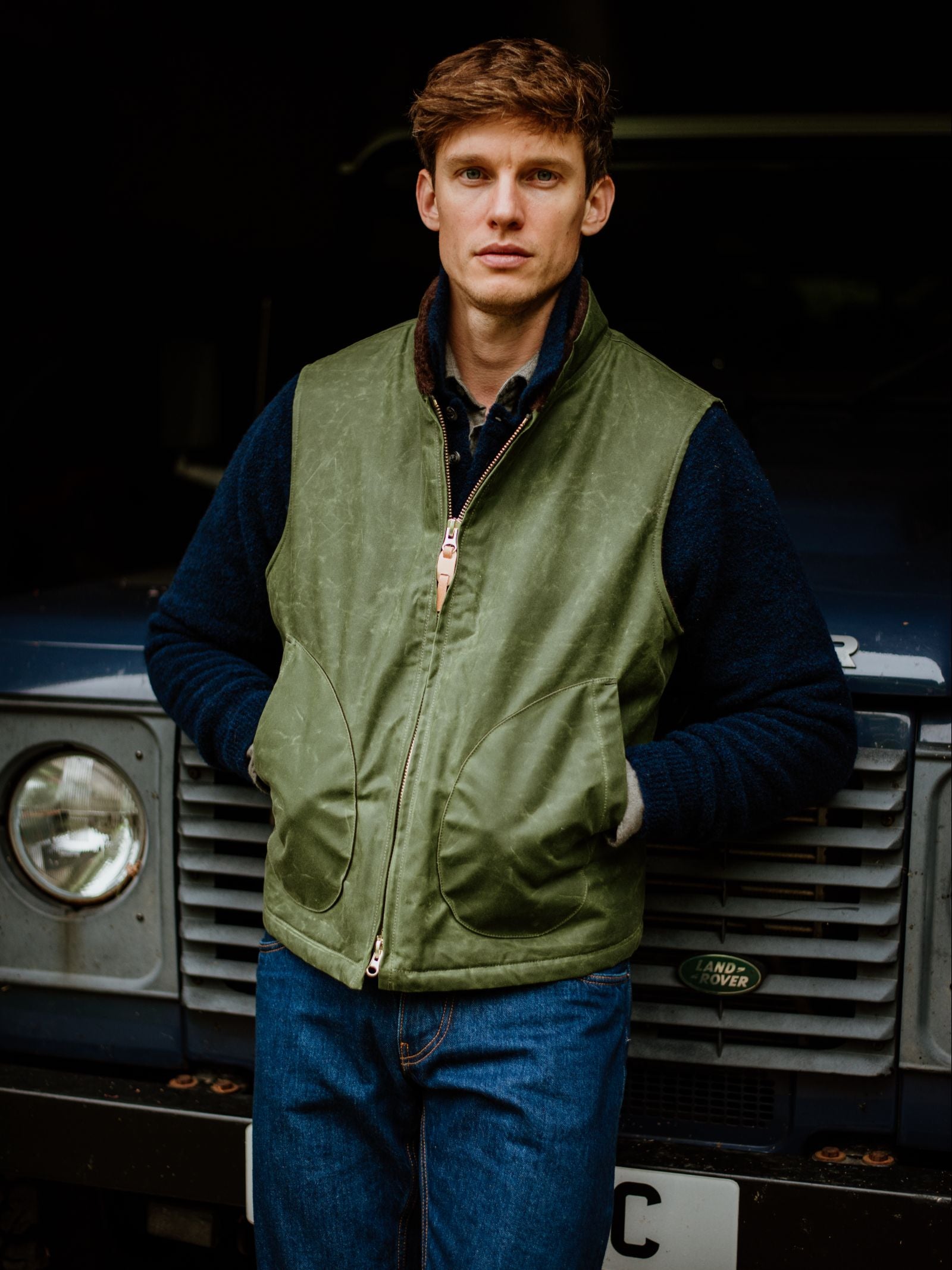 A young man wearing Campbell's of Beauly Wax Nehru Gilet and blue jeans stands in front of a dark Land Rover, looking at the camera with a neutral expression.