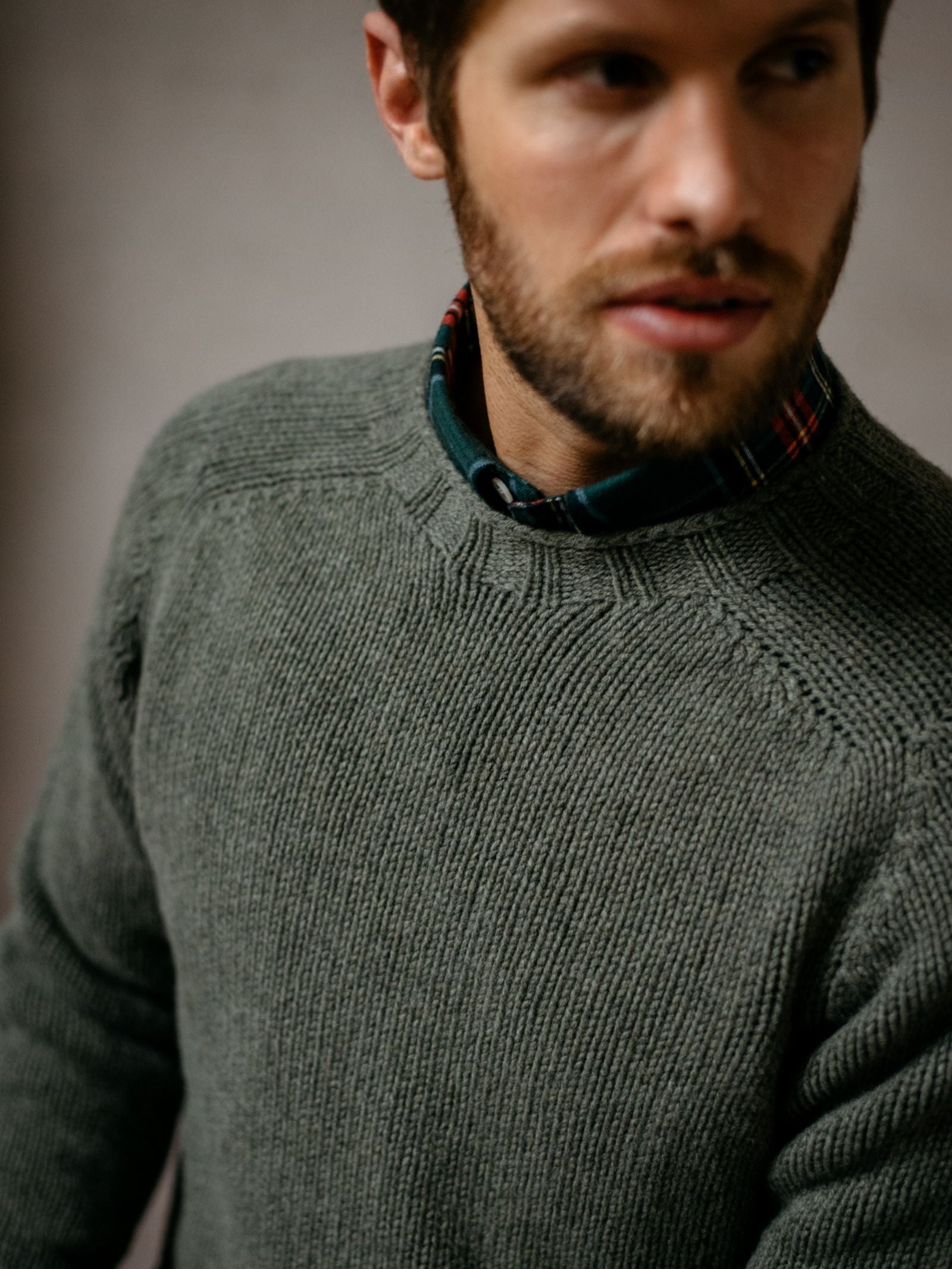 A bearded man wears the Campbells of Beauly Geelong Lambswool Chunky Crew Jumper in olive green over a dark green, red, and white plaid shirt. The softly focused image shows him from shoulders up against a neutral background.