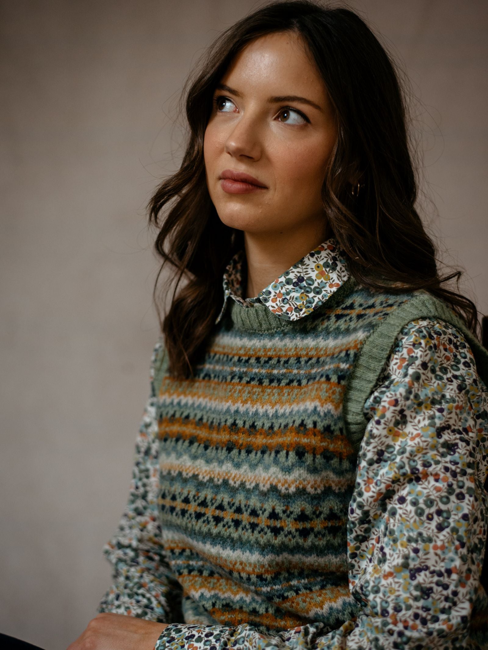 A woman with long brown hair gazes upward, wearing a Campbells of Beauly Liberty Print Cotton Shirt under a patterned sweater vest, seated against a softly lit neutral background.