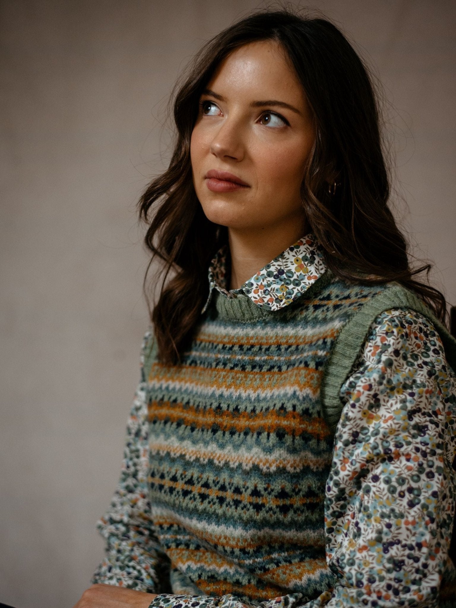 A young woman with wavy brown hair looks upward thoughtfully, wearing the Campbell's of Beauly Fairisle Crew Tank, knitted in Scotland from soft Shetland wool, layered over a floral button-up shirt against a neutral background.
