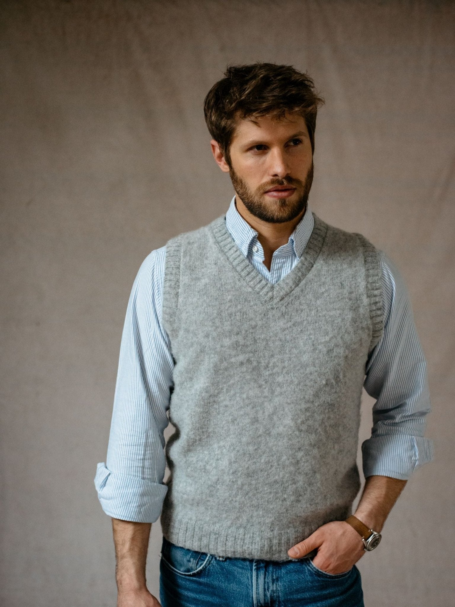 A man with brown hair and a beard wears a light blue striped shirt and the Campbells of Beauly Shetland Slipover grey V-neck knit vest, sleeves rolled up, standing with one hand in his pocket against a neutral background—a timeless classic.
