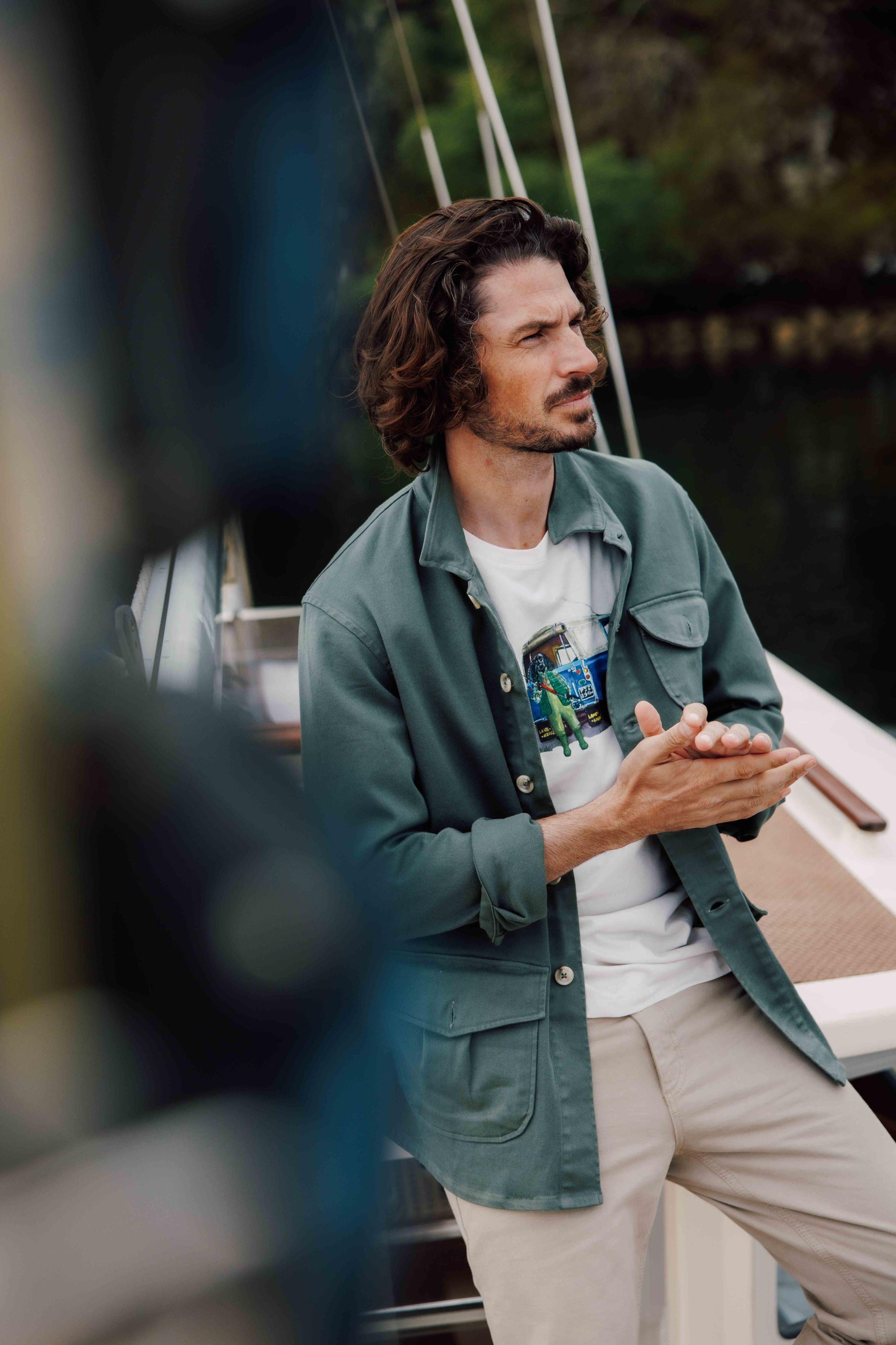 A man with wavy brown hair and a beard leans against a boat railing, wearing a green jacket over a graphic t-shirt and beige pants, looking thoughtfully into the distance.