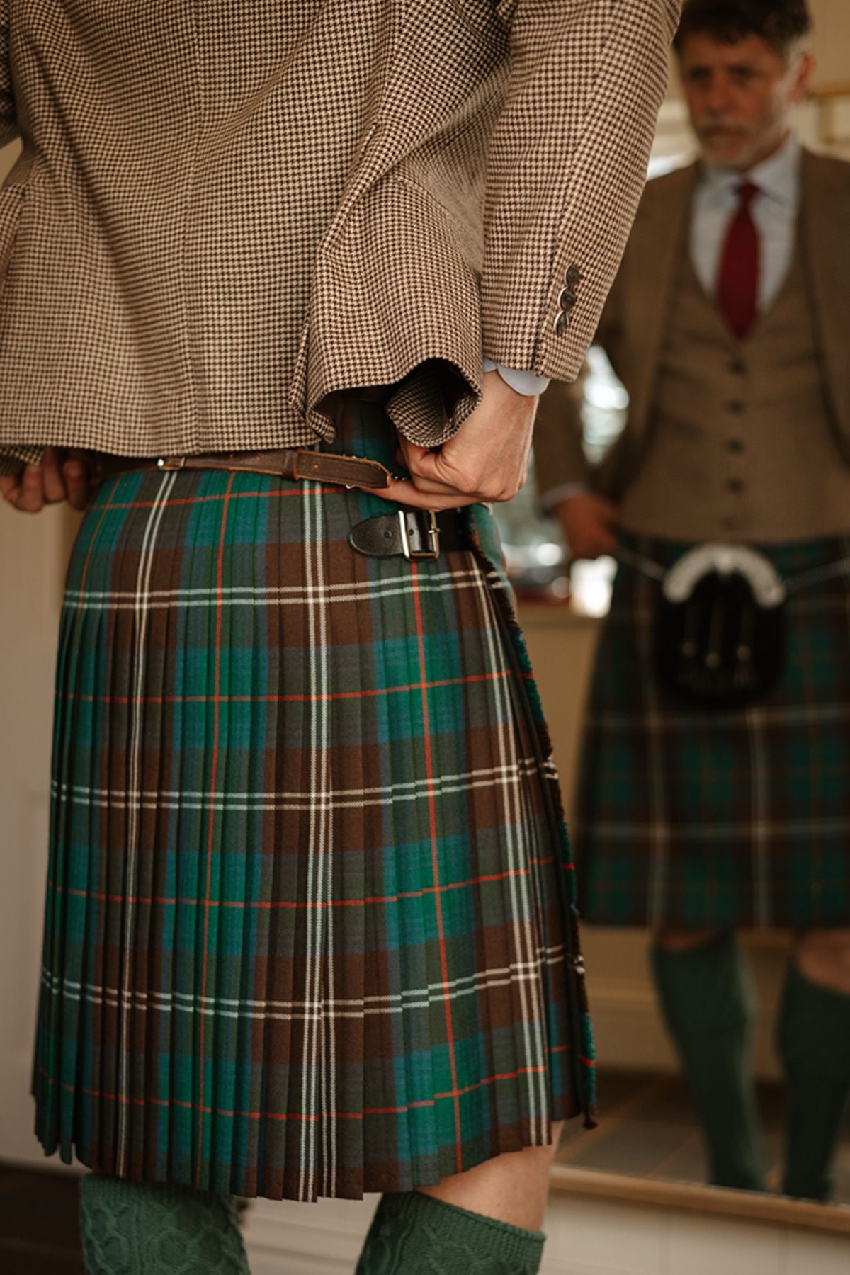 A man wearing a houndstooth jacket and a green and brown tartan kilt adjusts his belt in a mirror.