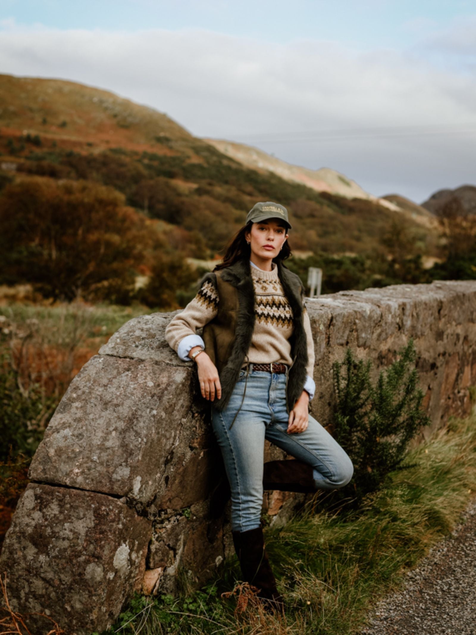 A woman in jeans, boots, a patterned sweater, a Campbell's of Beauly Auld Stock Sheepskin Reversible Gilet, and a cap leans against a stone wall in a grassy, hilly rural landscape with trees.