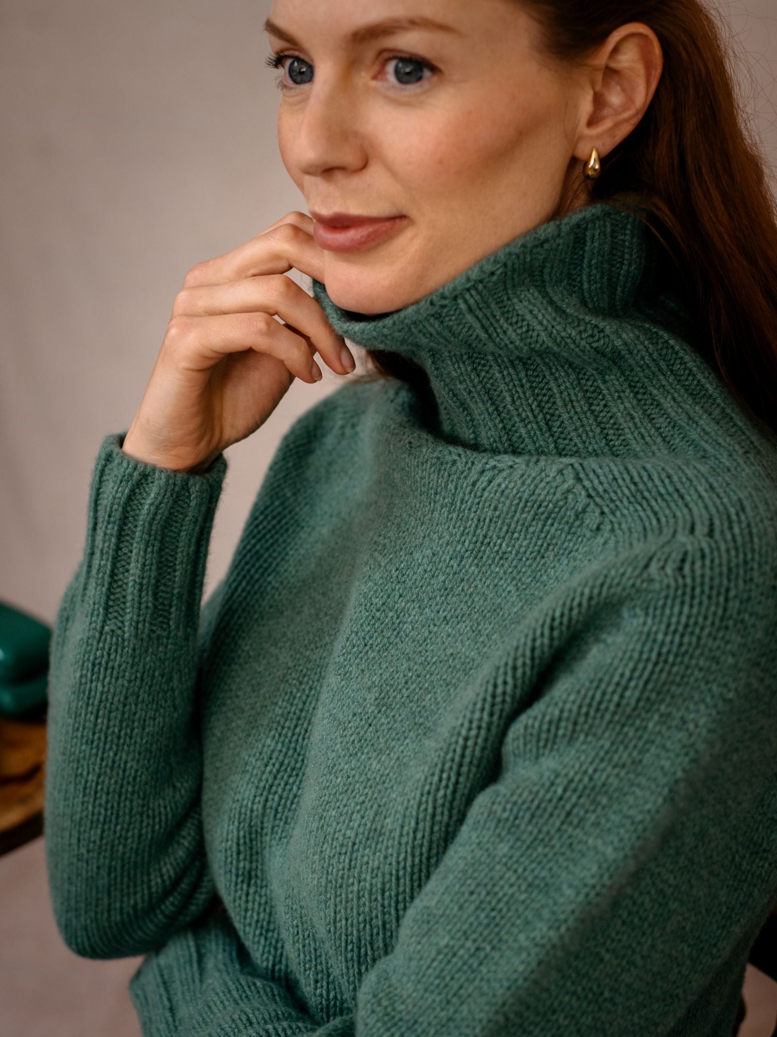 A woman with long brown hair, wearing a green Chunky Roll Neck Sweater by Campbells of Beauly and small gold hoop earrings, sits with her hand on her chin, looking thoughtful.