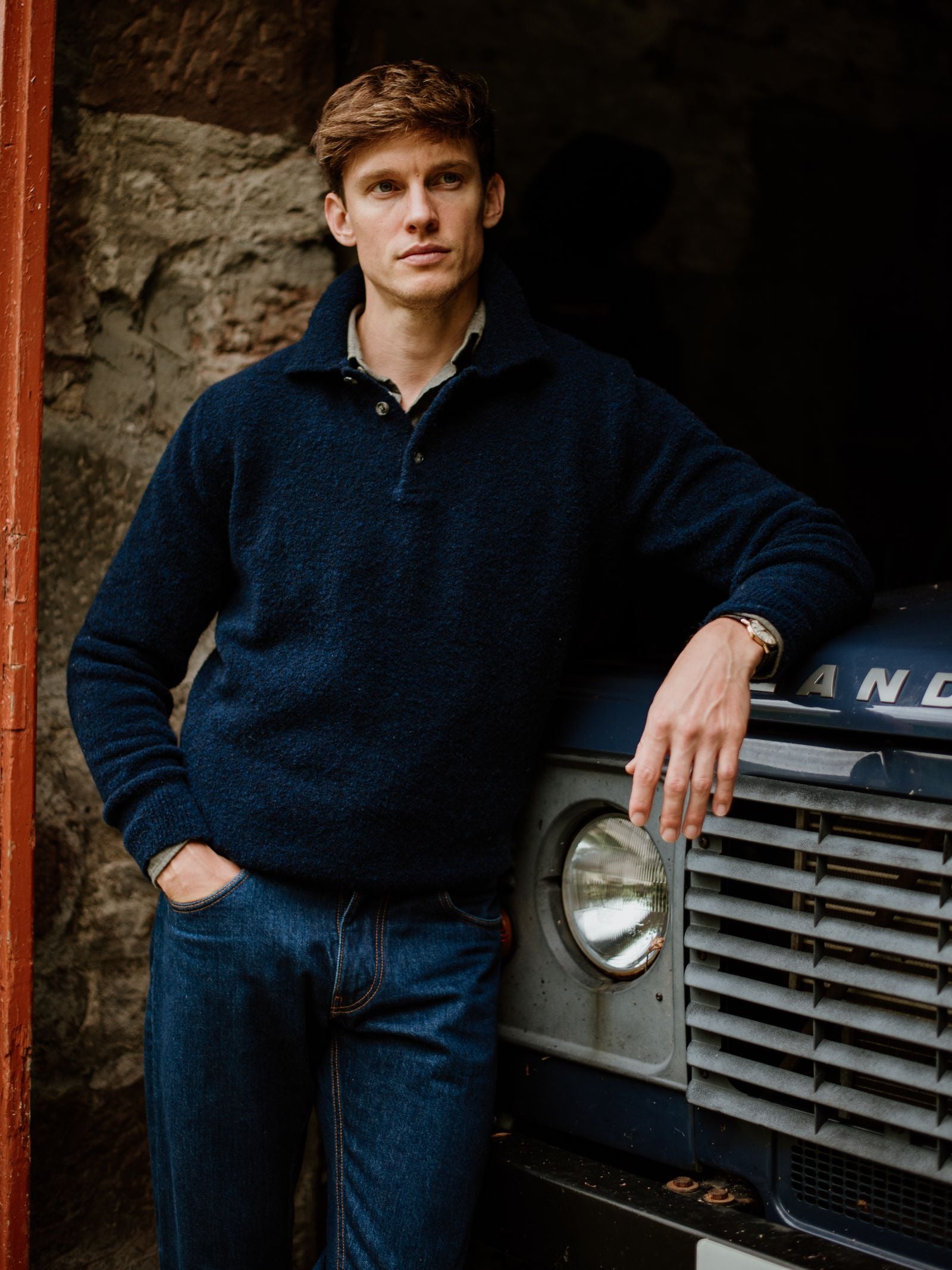 A man with short brown hair wearing the Campbell's of Beauly Auld Stock Bouclé Neck Jumper and jeans leans against a blue vehicle, one hand in his pocket and the other on the hood, looking off to the side.