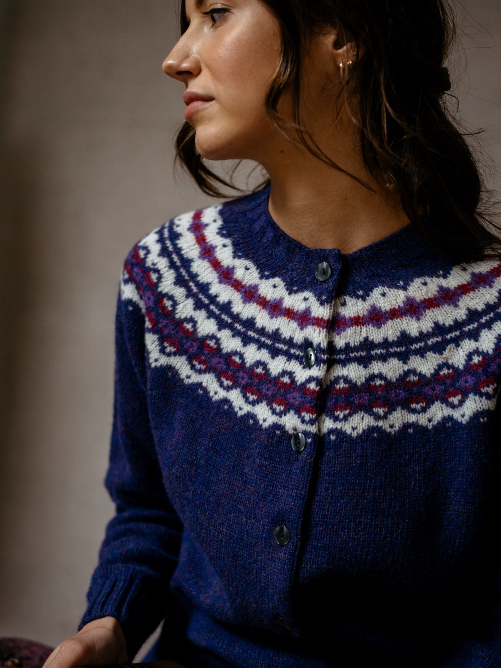 A woman with wavy brown hair wears the Campbells of Beauly Shetland Fairisle Cardigan in blue Pure New Wool, featuring a white, purple, and red yoke. Seated in soft light, she looks to the side—a classic Scottish knitwear piece.