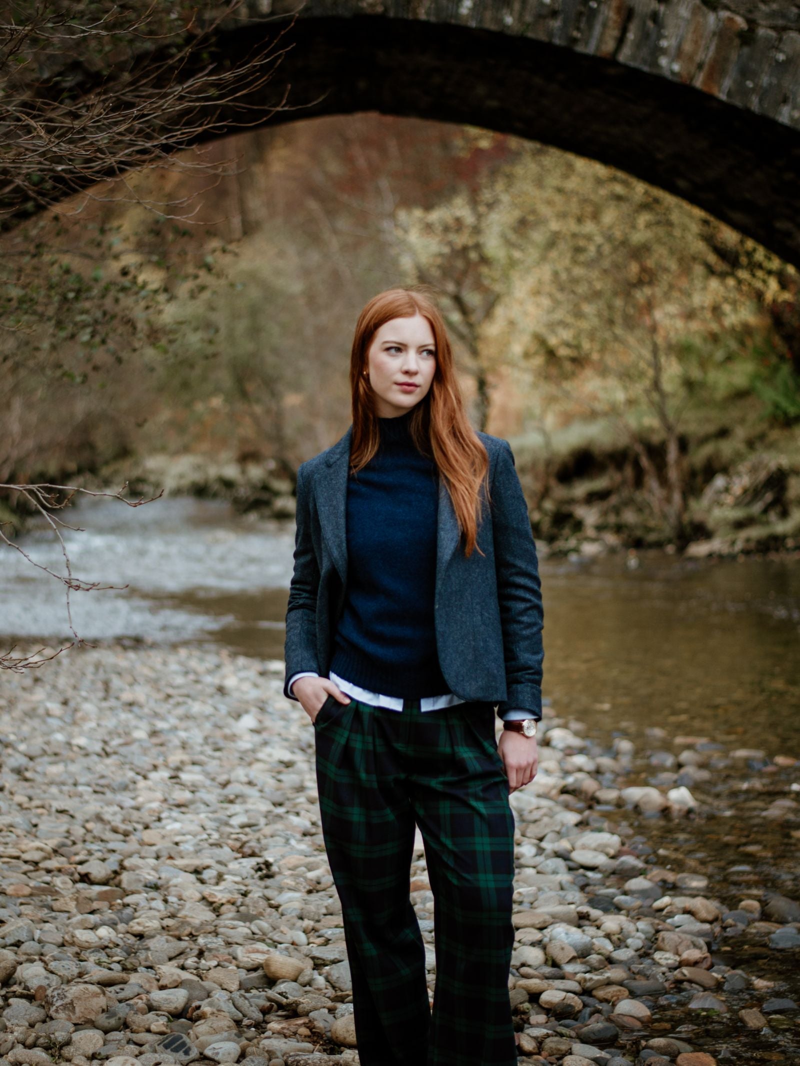 A woman with long red hair wears the House Jacket by Campbells of Beauly in navy on a rocky riverbank beneath a stone arch bridge, framed by trees and water—a portrait of timeless style.