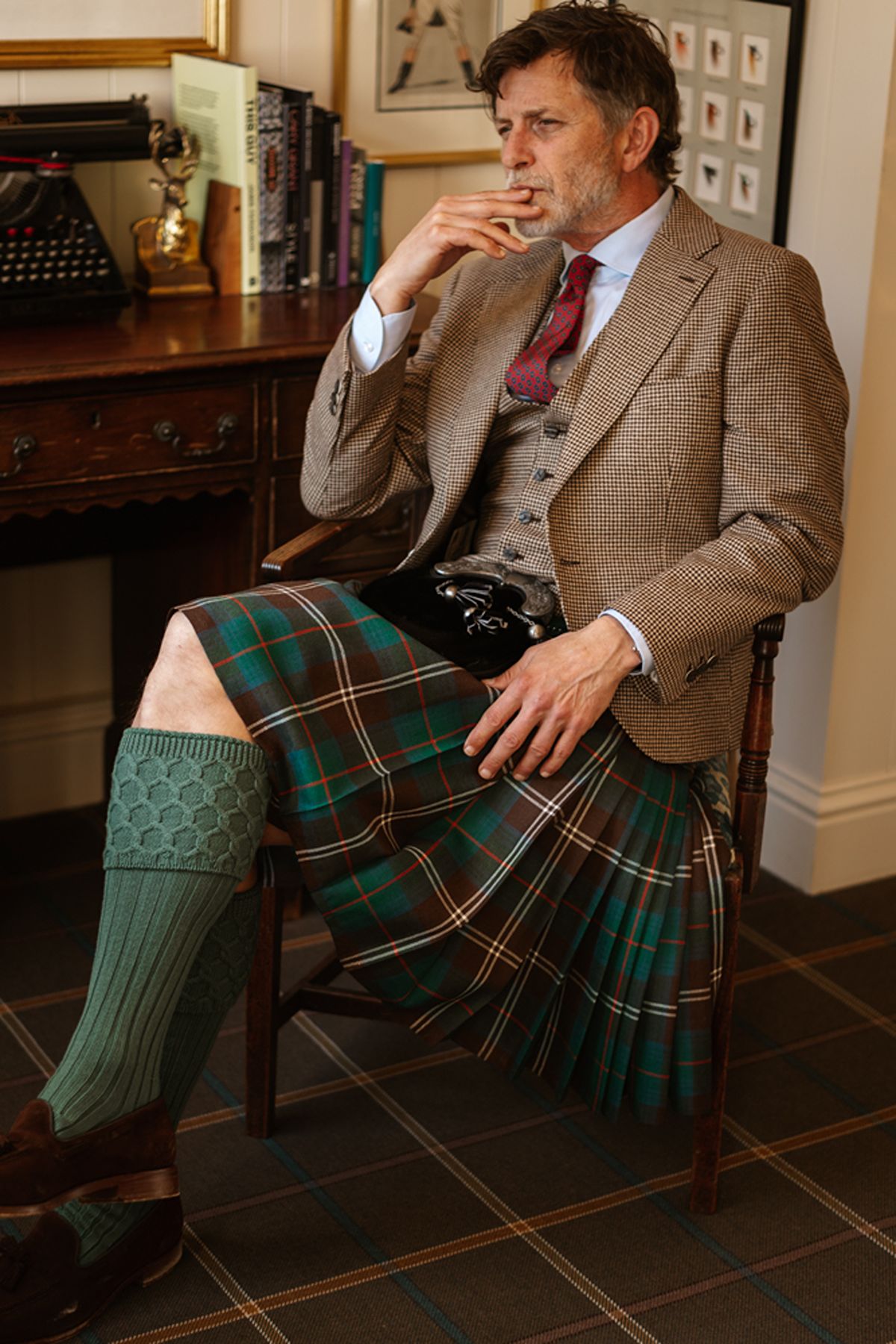 A man wearing a tweed jacket, waistcoat, red patterned tie, and tartan kilt sits in a wooden chair with his hand to his chin.