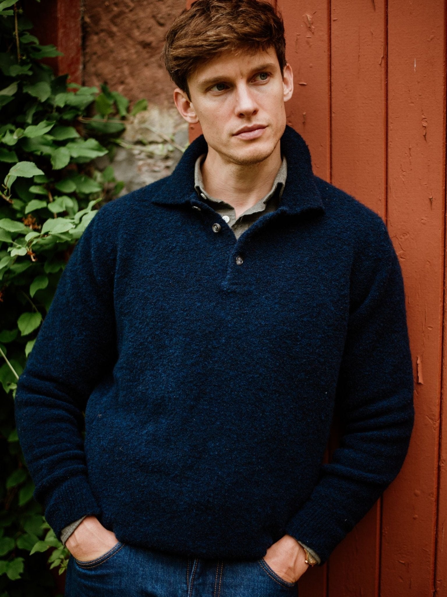 A man with short brown hair, wearing the Campbell's of Beauly Bouclé Polo Sweater and jeans, stands with hands in his pockets before a rustic red wooden wall and green foliage, gazing thoughtfully to the side.