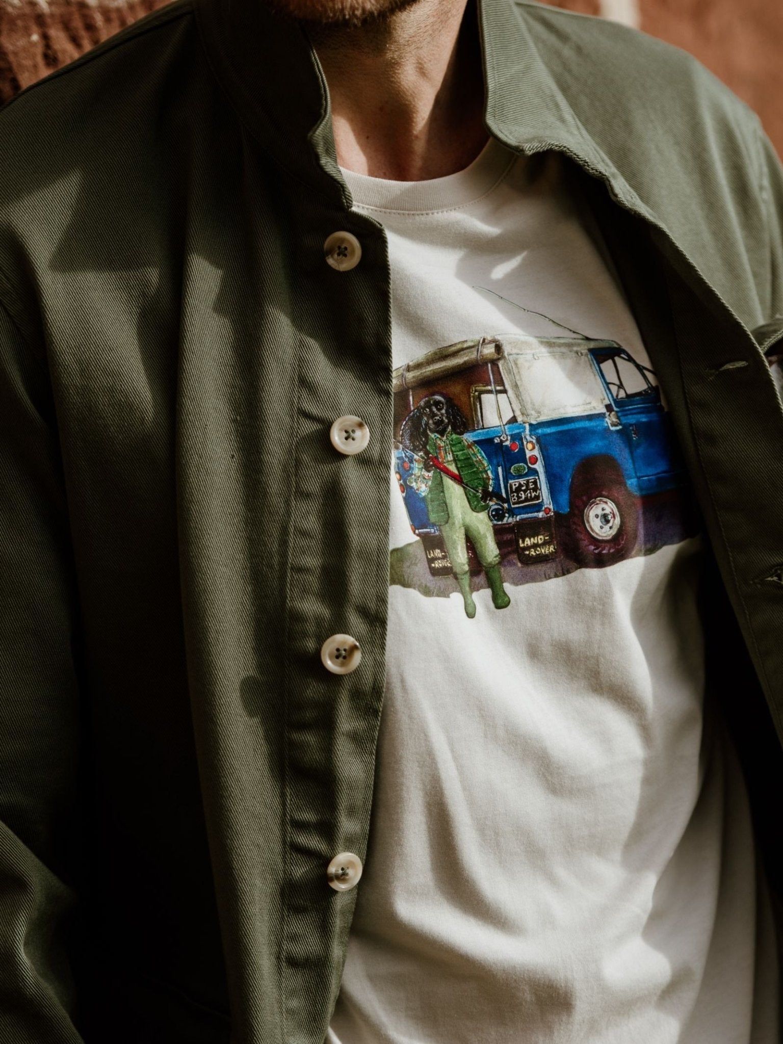 A person wearing an olive green jacket over a Campbells of Beauly Land Rover Cocker T-Shirt, which is white cotton and features a graphic of a blue Land Rover with a person standing in front. Only the upper torso is visible.
