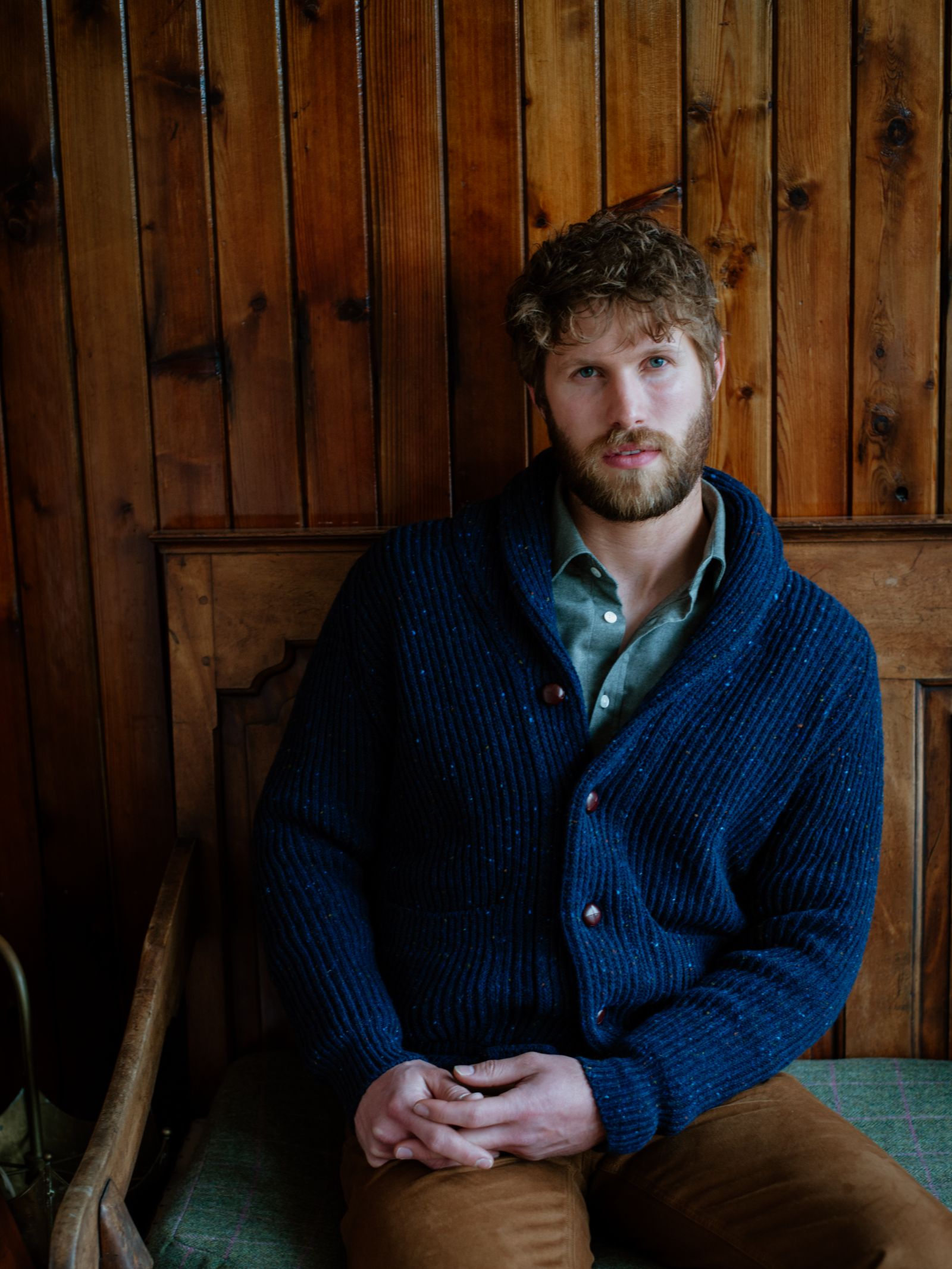 A bearded man with wavy brown hair sits on a wooden bench against a wood-paneled wall, wearing Campbells of Beauly Donegal Lambswool Shawl Collar Cardigan with leather buttons over a green shirt and brown pants, gazing thoughtfully at the camera.