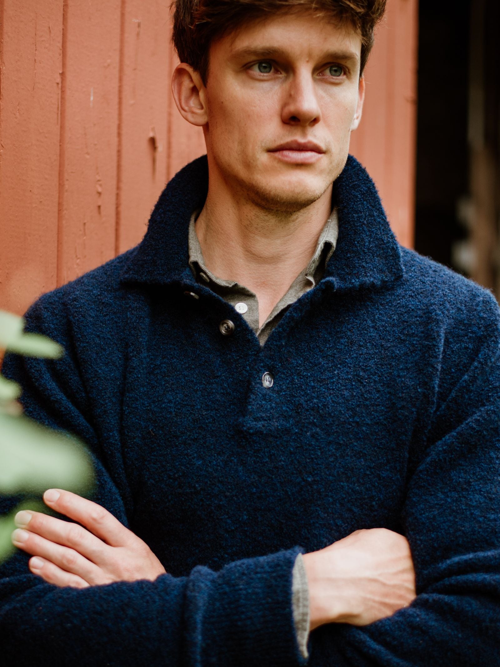 A young man with light brown hair, wearing the Campbell's of Beauly Bouclé Polo Sweater in dark blue, stands with arms crossed before a reddish-brown wooden wall, gazing thoughtfully into the distance.
