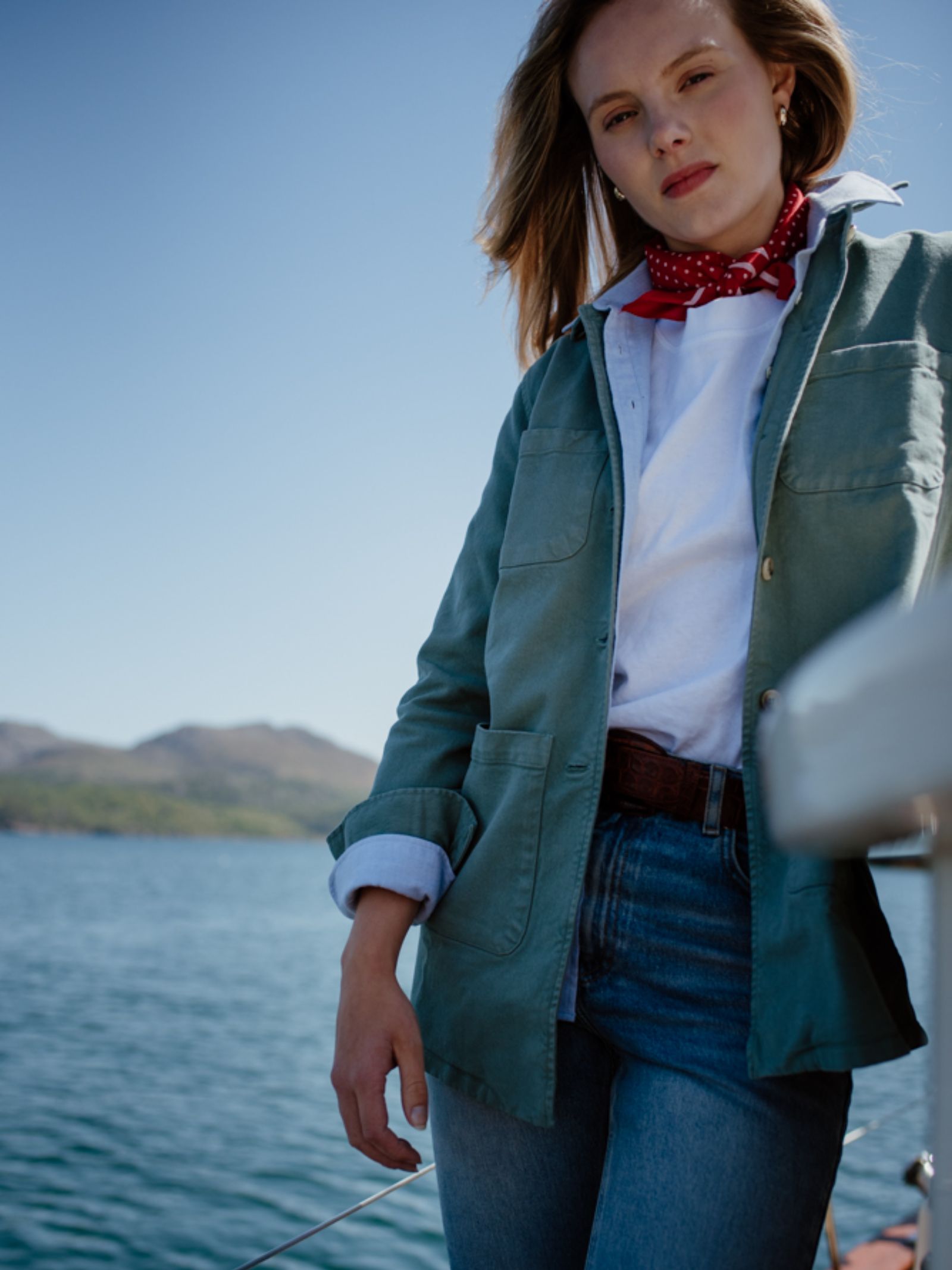 A woman stands on a boat by the water, wearing Campbells of Beaulys green Summer Chore Jacket with a relaxed fit, paired with a white shirt, red scarf, and jeans. Mountains and blue sky form the backdrop.