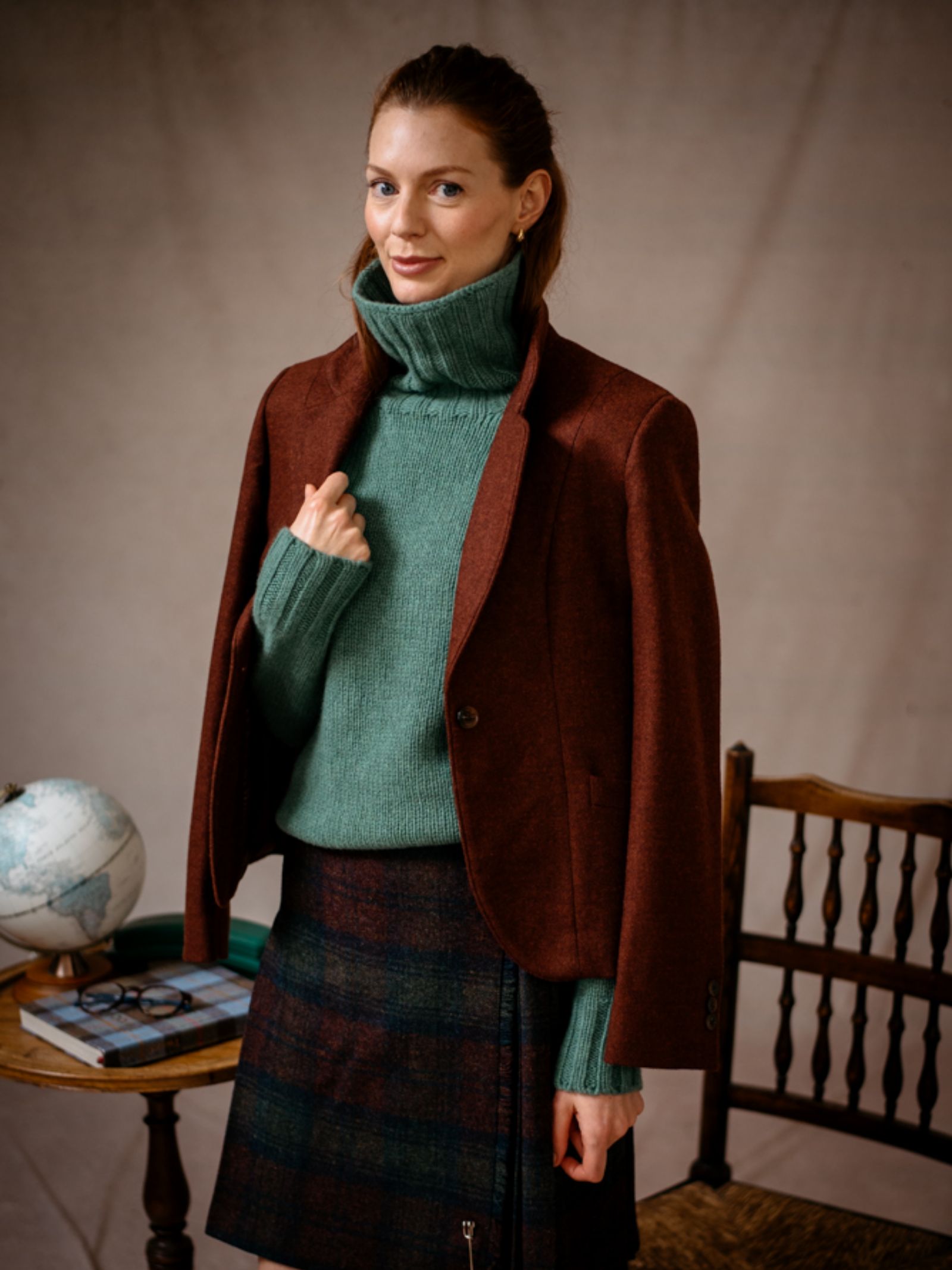 A woman stands indoors in a green turtleneck and brown blazer, wearing the Campbells of Beauly Tweed Kilt. She is beside a table with a globe, book, and glasses, with a wooden chair behind her.