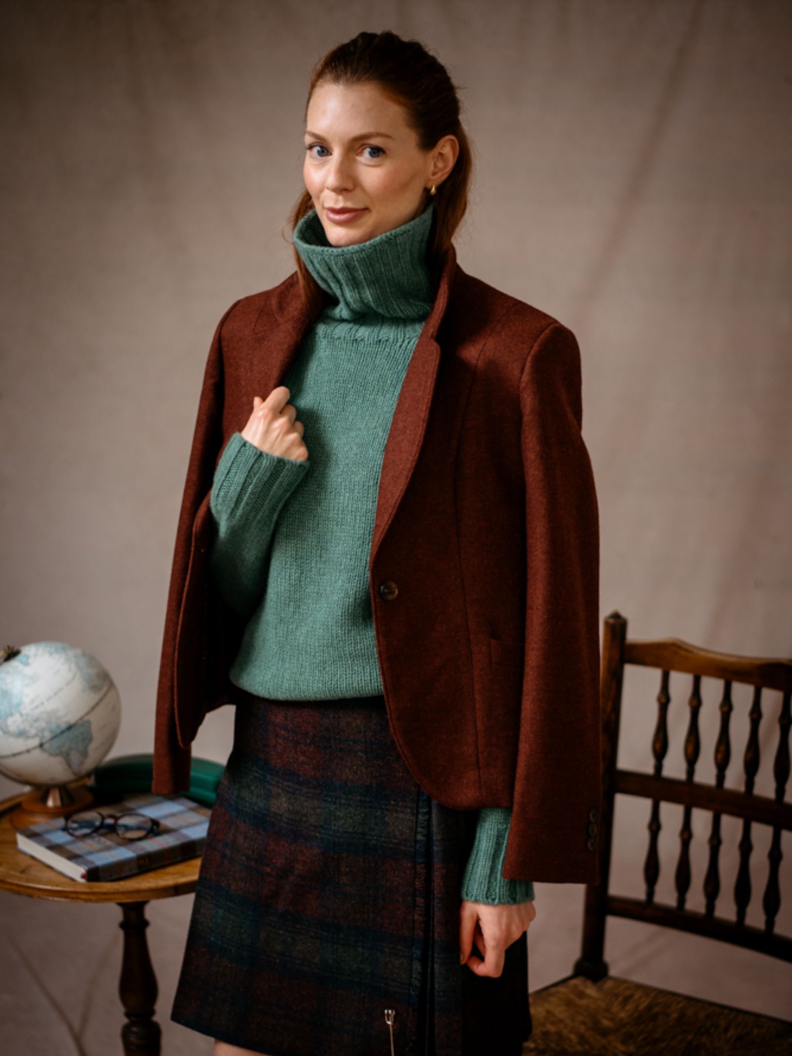 A woman stands indoors beside a table with a globe and books, draped in the Campbells of Beauly House Jacket over her green turtleneck sweater, exuding timeless style.
