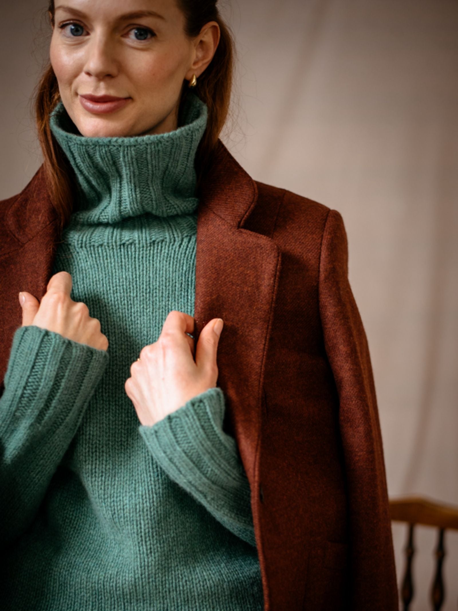 A woman with fair skin and red hair wears the Campbell’s of Beauly House Jacket in classic herringbone tweed over a thick, light green turtleneck, holding the lapels and smiling softly at the camera against a neutral background.