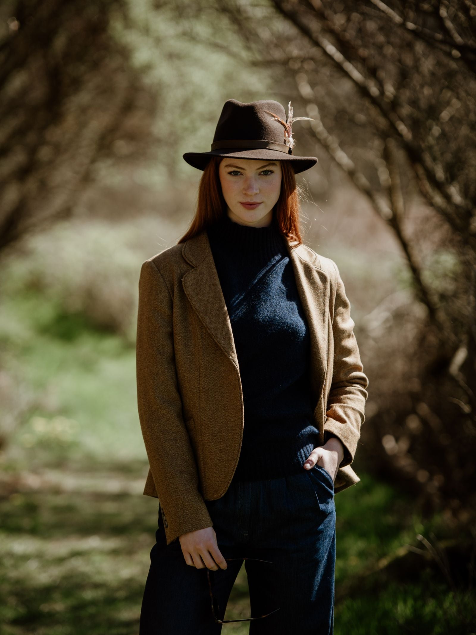 A woman with long red hair stands on a sunlit green path, wearing the Hicks & Brown Gamebird Fedora with a feather wrap, a brown blazer, dark sweater, and blue pants, one hand in her pocket among the trees.