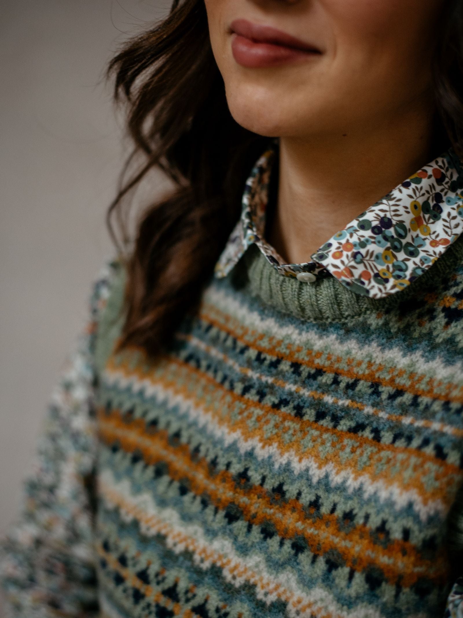 A woman with wavy brown hair wears a floral collared shirt under the Campbell's of Beauly Fairisle Crew Tank, a multicolored patterned knit crafted in Scotland. The lower half of her face shows a slight smile.