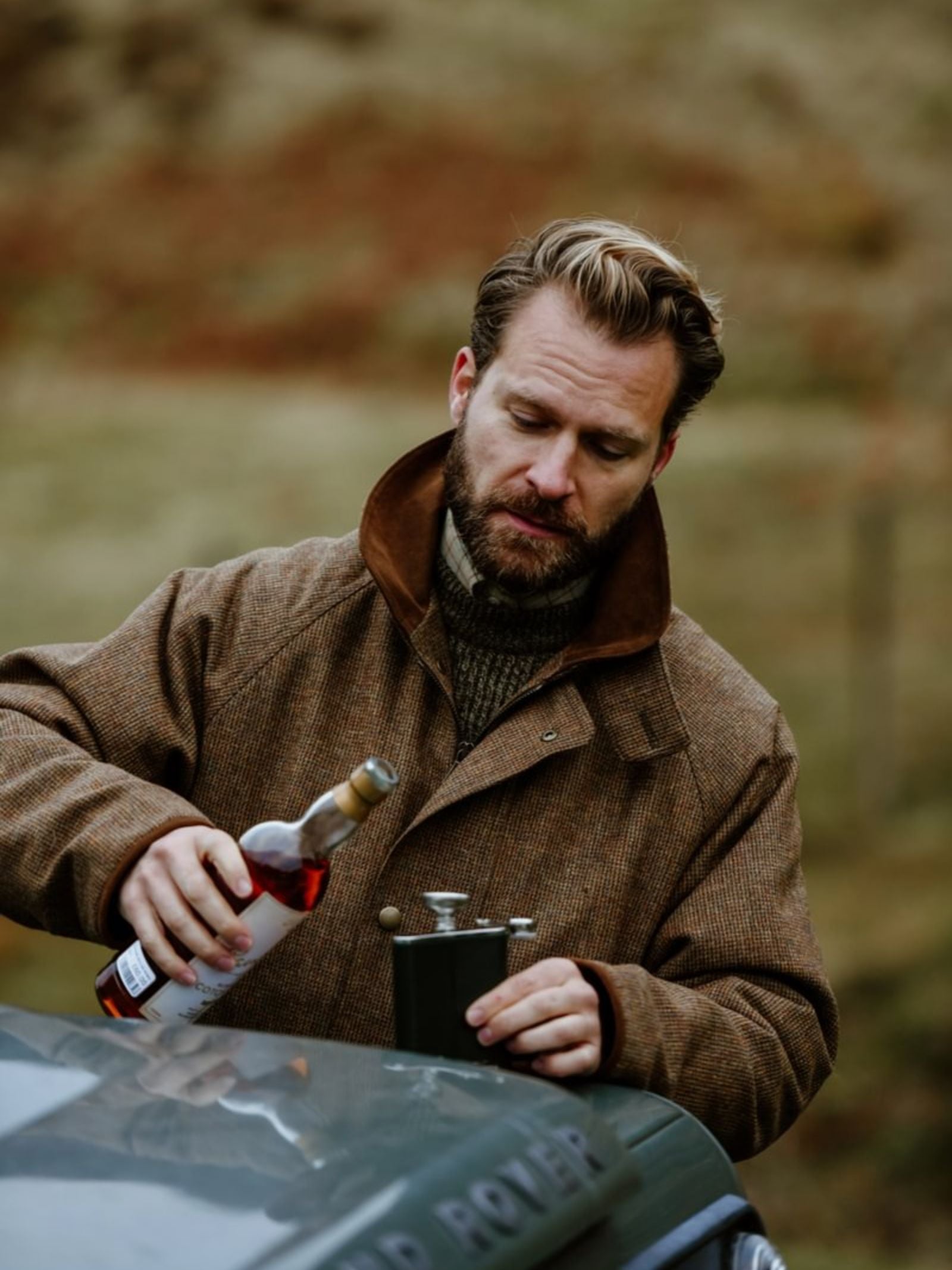 A bearded man in a brown coat pours a drink into the Campbell's of Beauly x Ettinger 6oz Forest Green Hip Flask beside a green vehicle outdoors, with grassy, blurred scenery in the background.