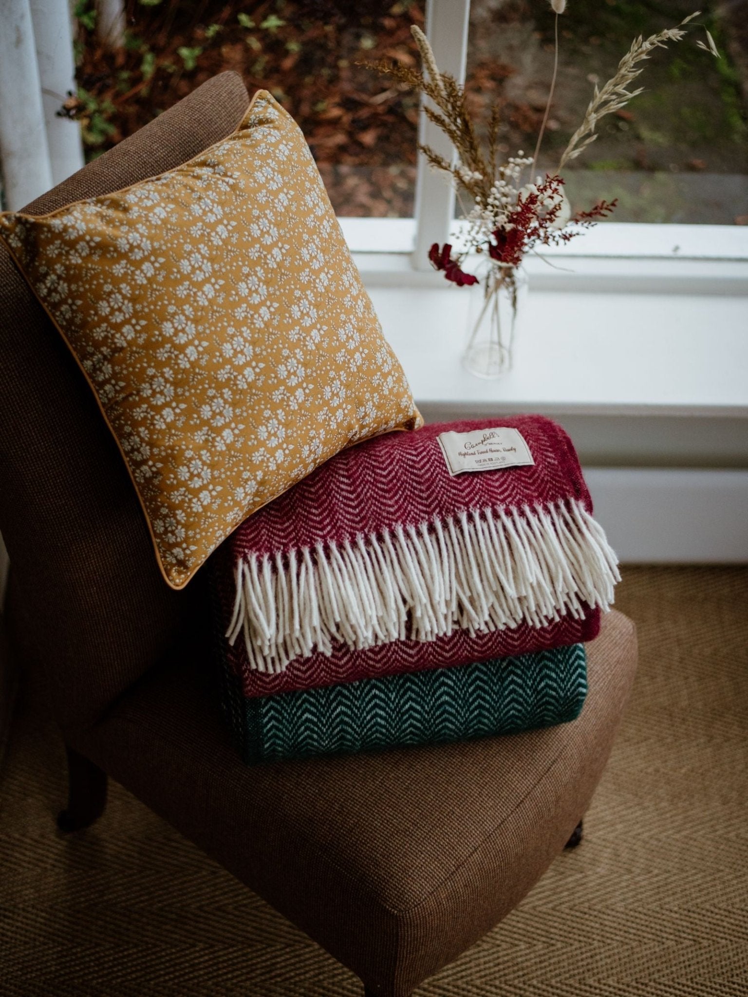 A brown chair holds a mustard yellow floral pillow and two folded woven blankets, one maroon and one green, both with white fringes. Nearby, Liberty Print Cushion Covers by Campbells of Beauly and a glass vase with dried flowers sit on the windowsill.