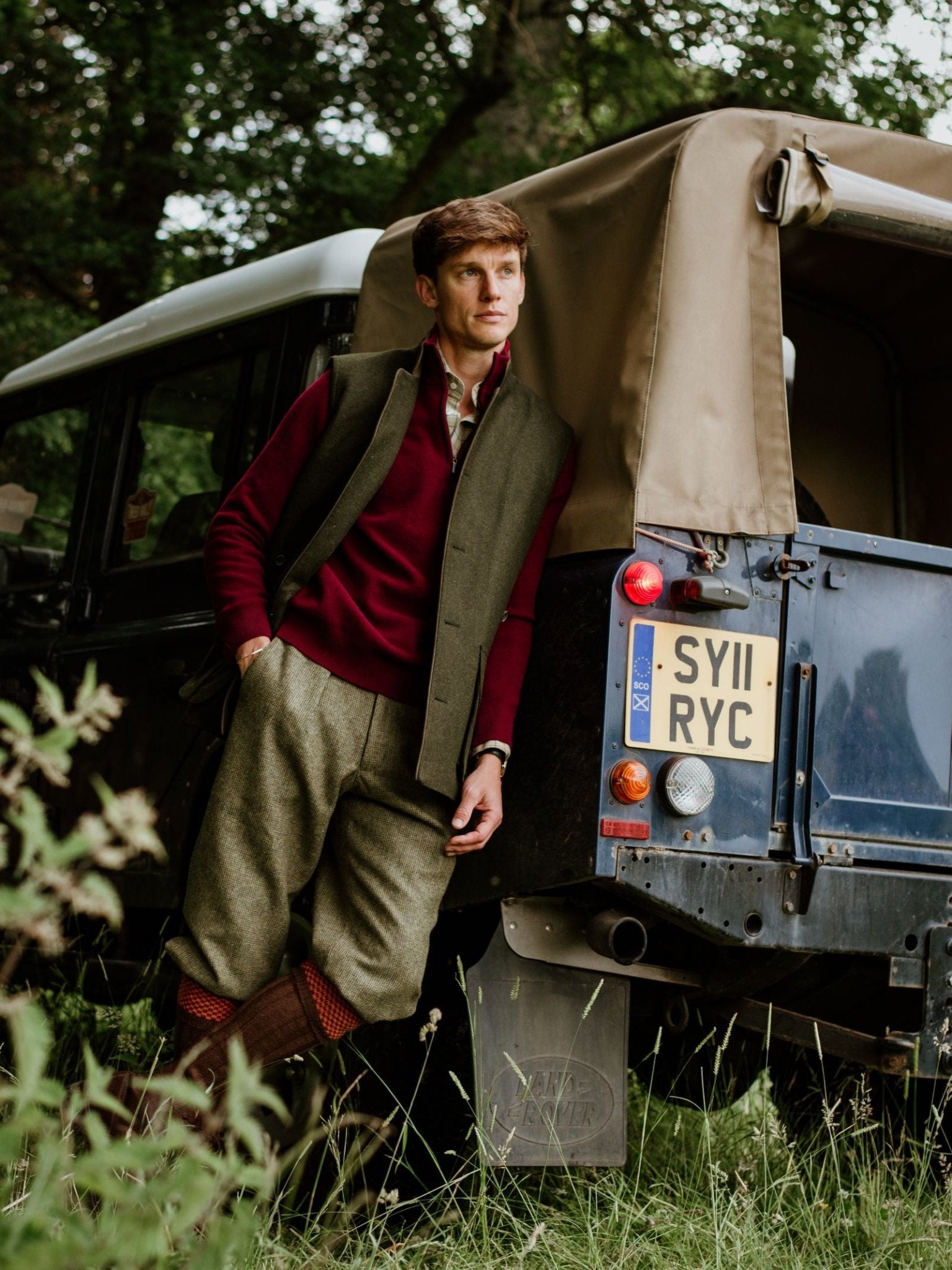 A man in classic country attire leans against a vintage off-road vehicle. He wears a red sweater, grey vest, tweed trousers, brown boots, and Hopstitch Shooting Socks from Campbell’s of Beauly—perfect fieldwear amidst the greenery.