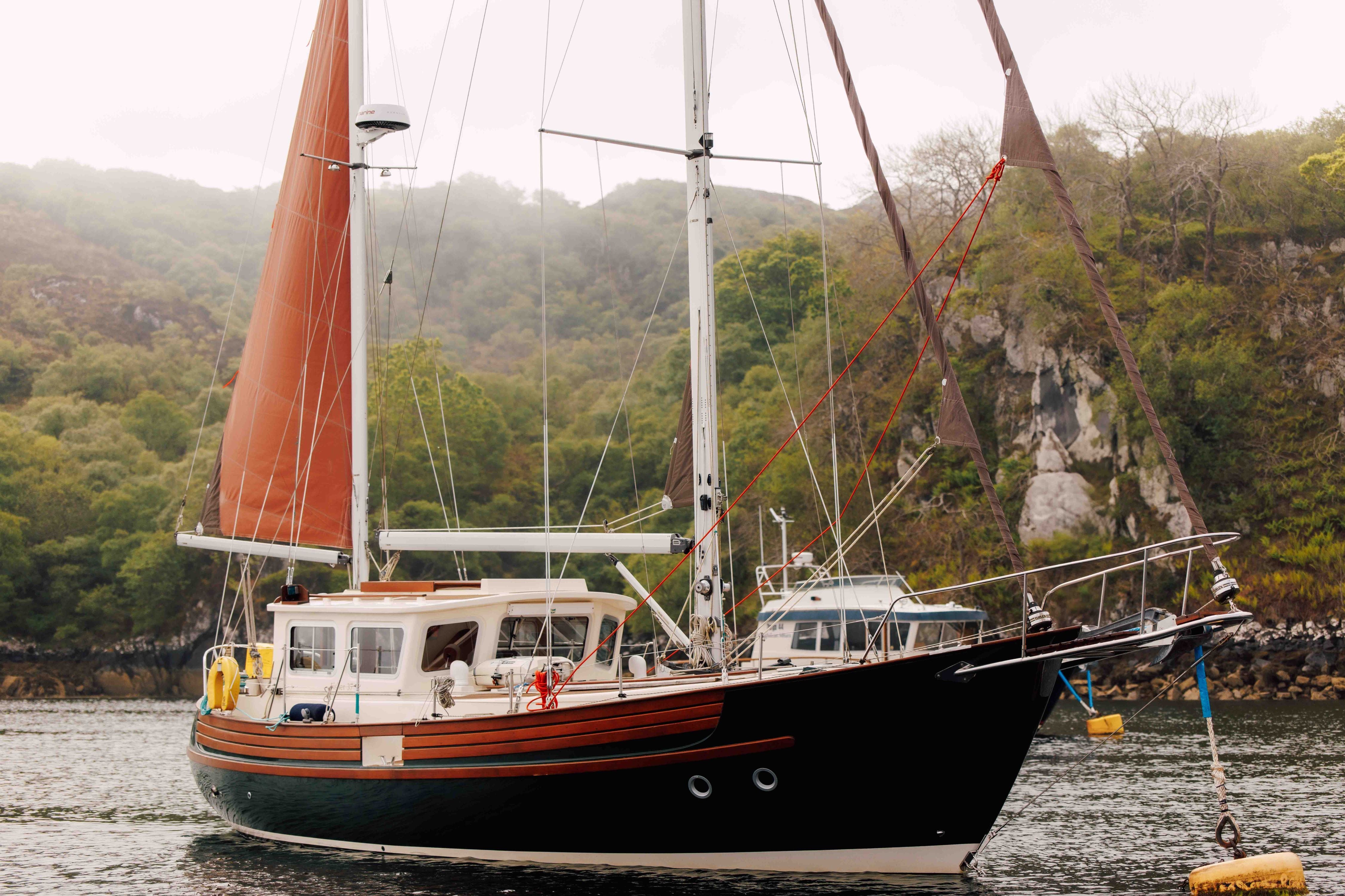 A black sailboat with reddish-brown sails is anchored on calm water near a forested, rocky shoreline. The boat has a white cabin and wooden accents, and another vessel is partially visible in the background.