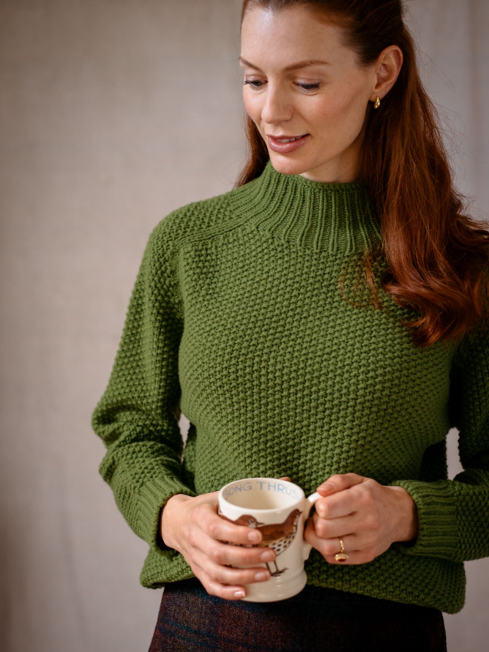 A woman in the Chunky Moss Stitch Polo by Campbells of Beauly holds a ceramic mug featuring an owl and the words LONG THRUSH. She has long brown hair, gold earrings, and looks down with a slight smile.