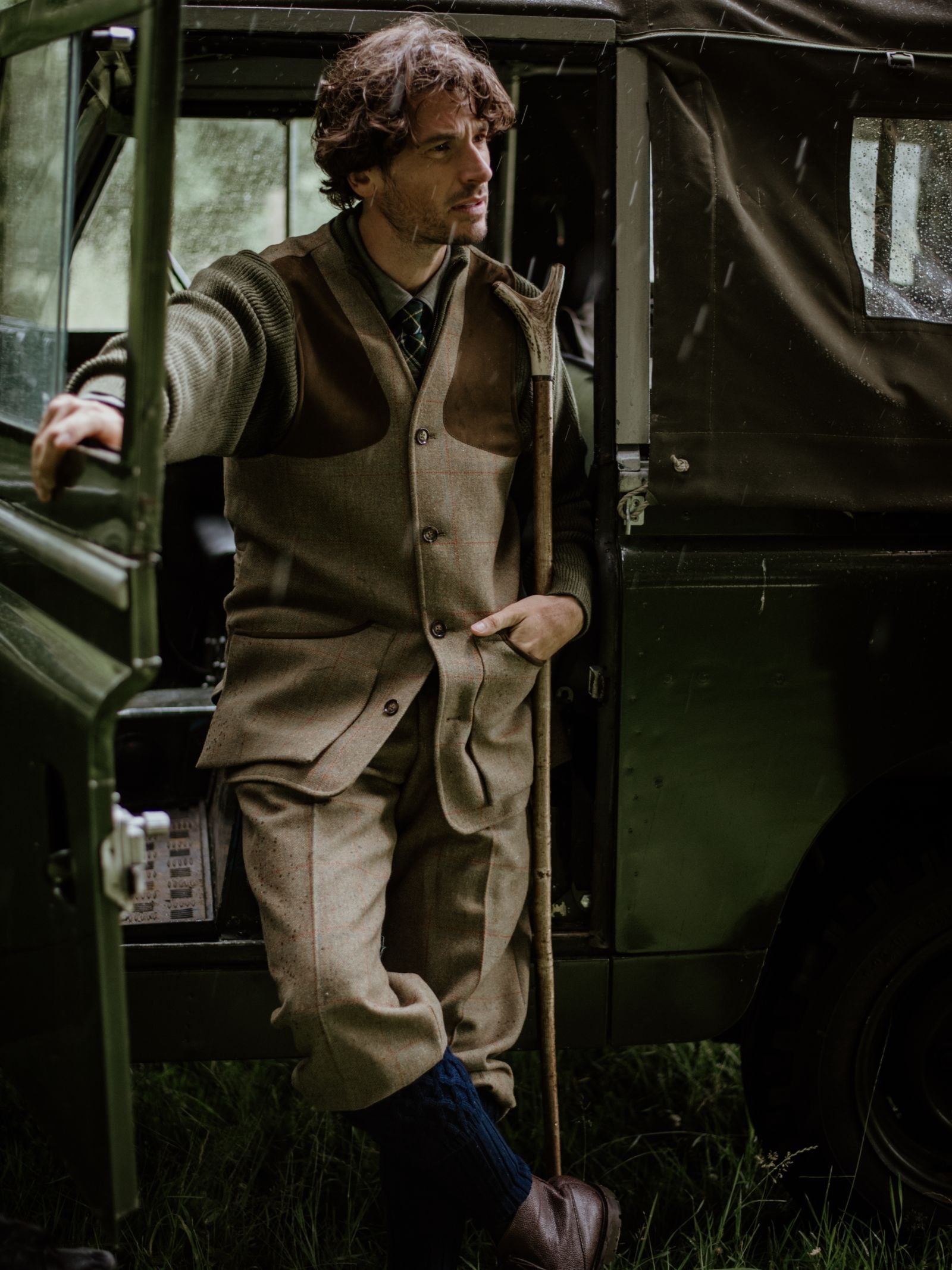 A man in traditional tweed hunting attire and a Campbell’s of Beauly Tweed Shooting Waistcoat leans against a dark green Land Rover with a wooden walking stick, grass beneath the vehicle.