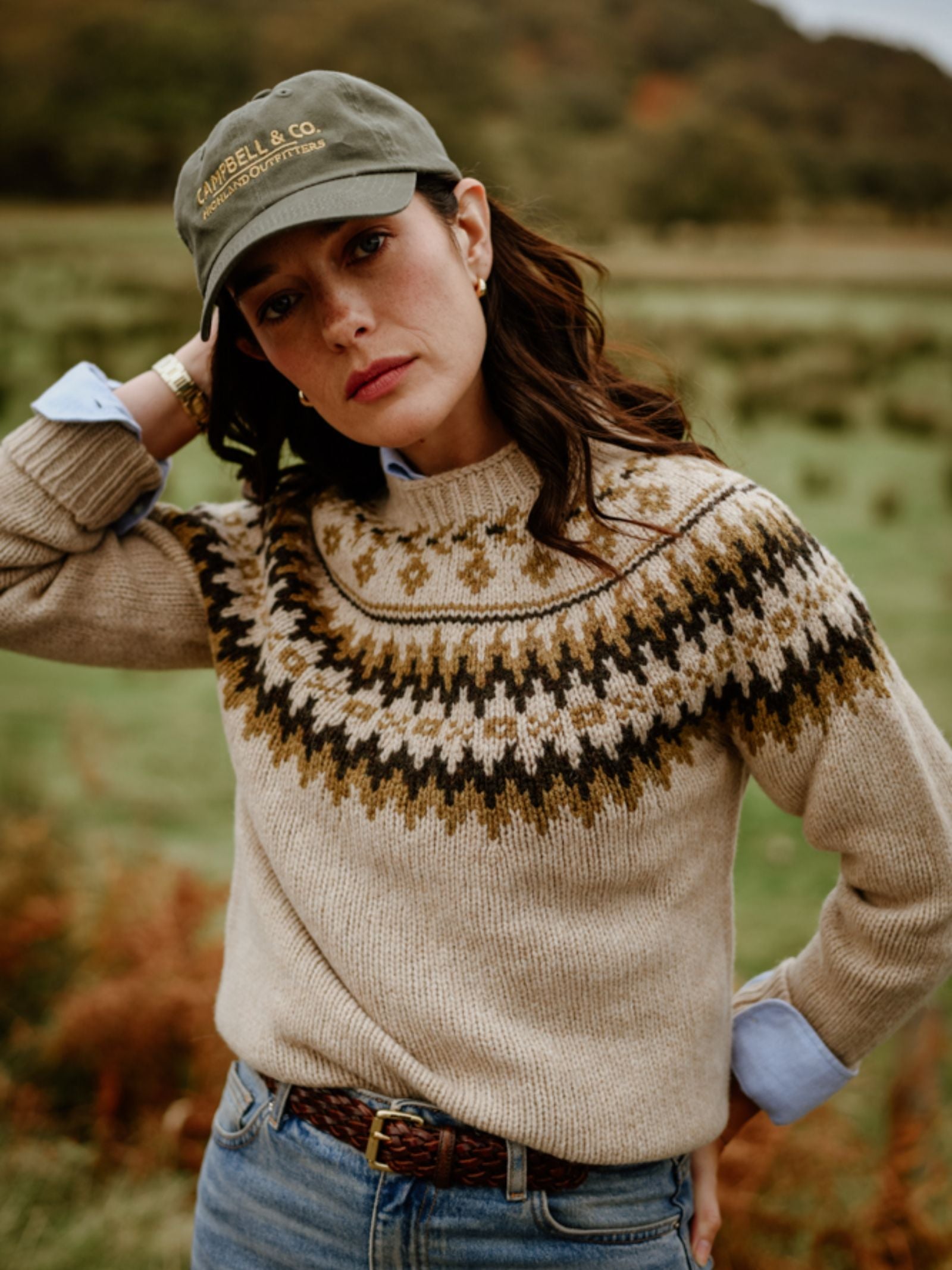 A woman in a Campbell's of Beauly Chunky Fairisle Jumper, blue jeans, and a green cap stands on grass with trees and hills behind her, one hand on her head, looking at the camera.