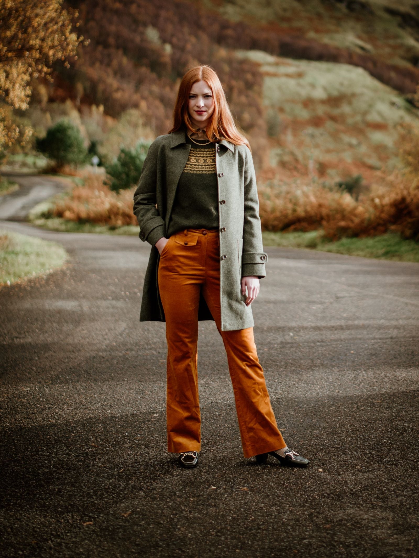 A woman with long red hair stands on a paved road, wearing Campbell’s of Beauly’s Auld Stock Heath Collar Coat in olive green, styled with a patterned sweater and orange pants. A hilly autumn landscape sets an inviting everyday wear scene.