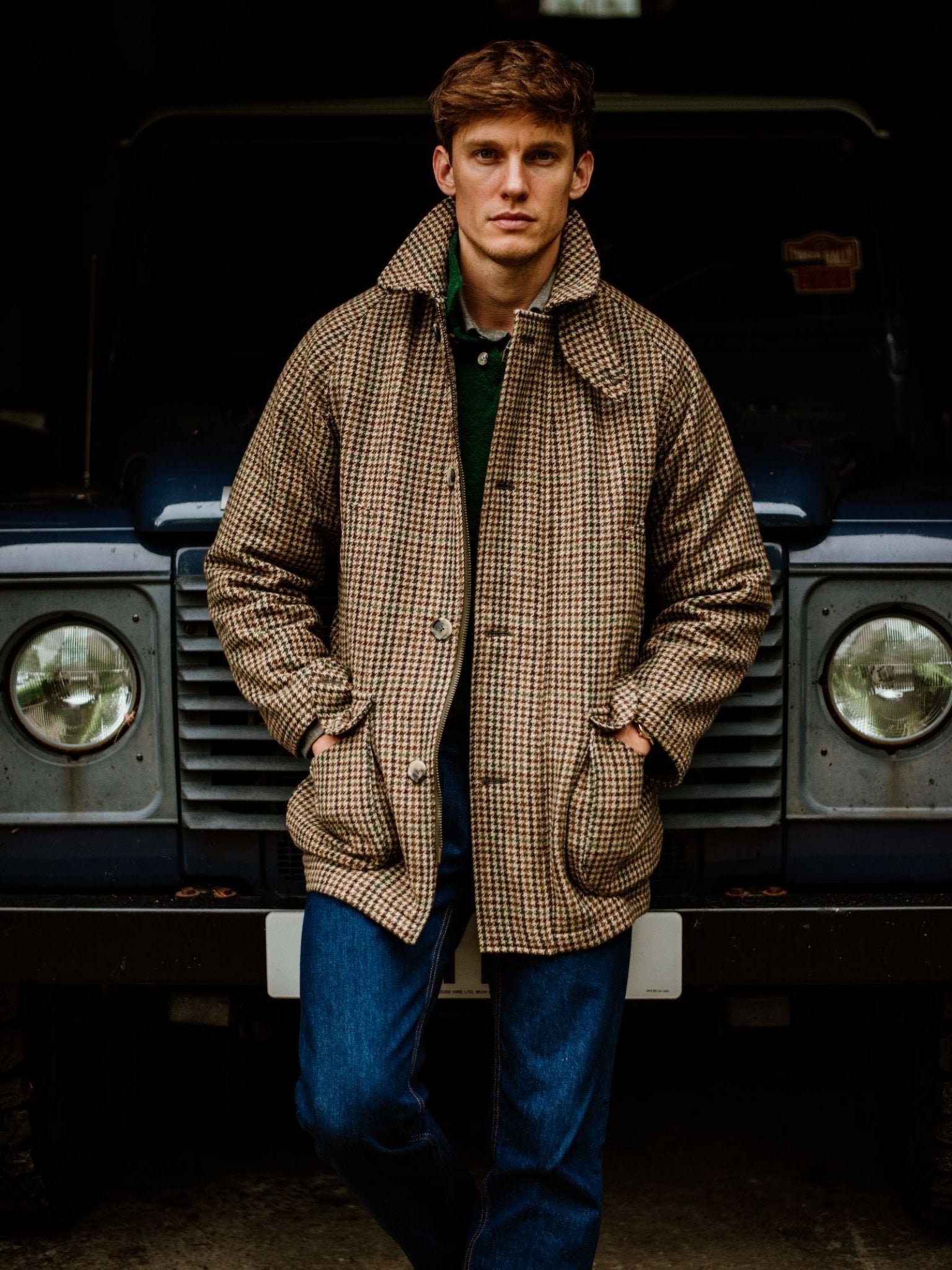 A man in a Cannich Coat by Campbell's of Beauly, crafted from Austrian wool, stands before a dark car with round headlights, hands in his pockets, embodying the spirit of country life as he looks at the camera.