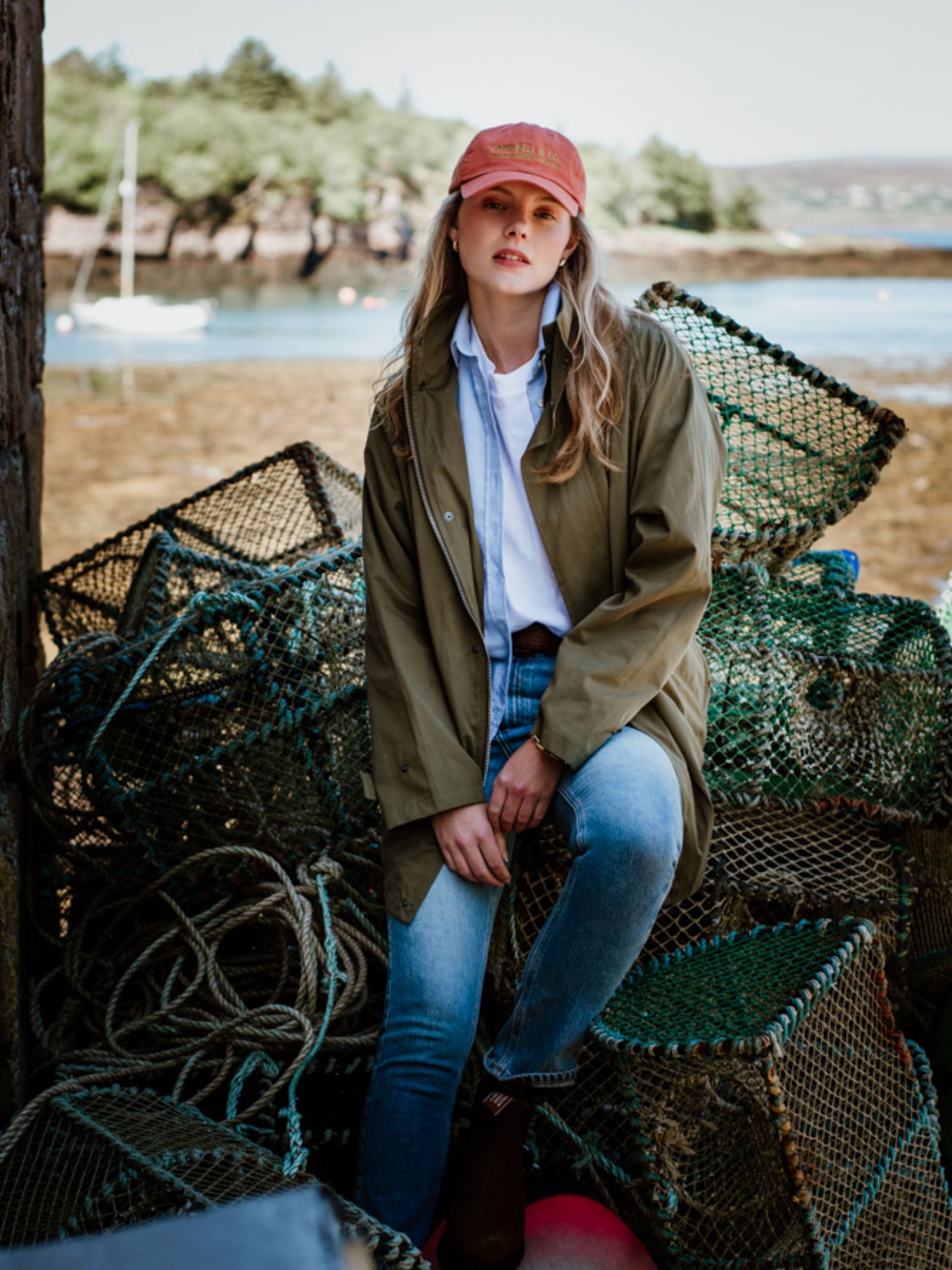 Wearing the Campbells Baseball Cap by Campbells of Beauly, a woman in a green jacket and jeans sits on fishing nets and lobster traps by the water, blending comfort and style with trees and a boat in the background.