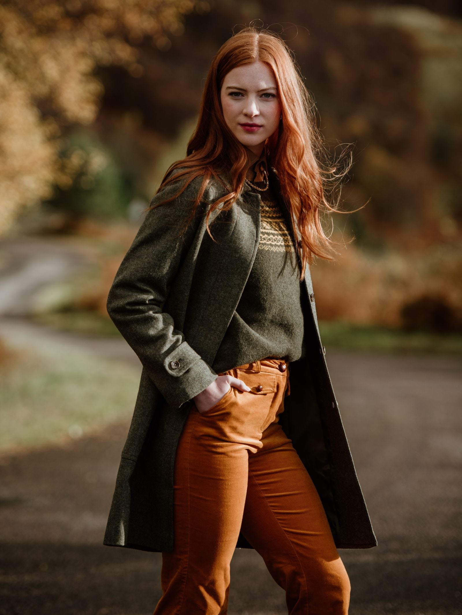 A woman with long red hair stands outdoors on a path, wearing the Campbells of Beauly Heath Collar Coat, a patterned sweater, and rust-colored pants. Autumn trees and soft sunlight create a warm, inviting backdrop for everyday style.