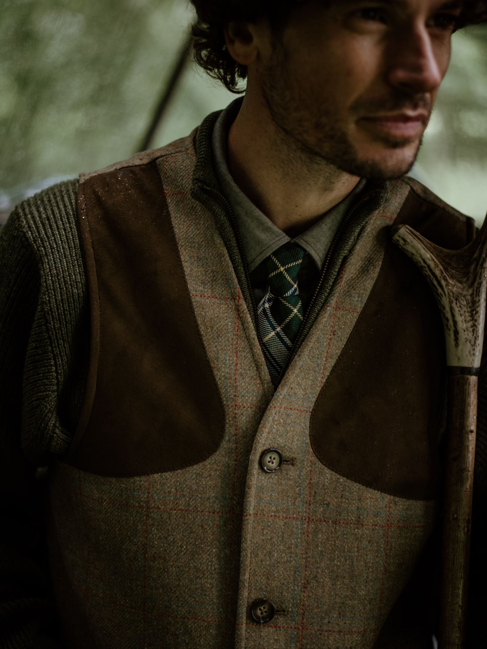 A man in a Campbell's of Beauly Tweed Shooting Waistcoat holds an antler. His face is partly visible against a blurred forest, capturing the timeless style of countryside pursuits.
