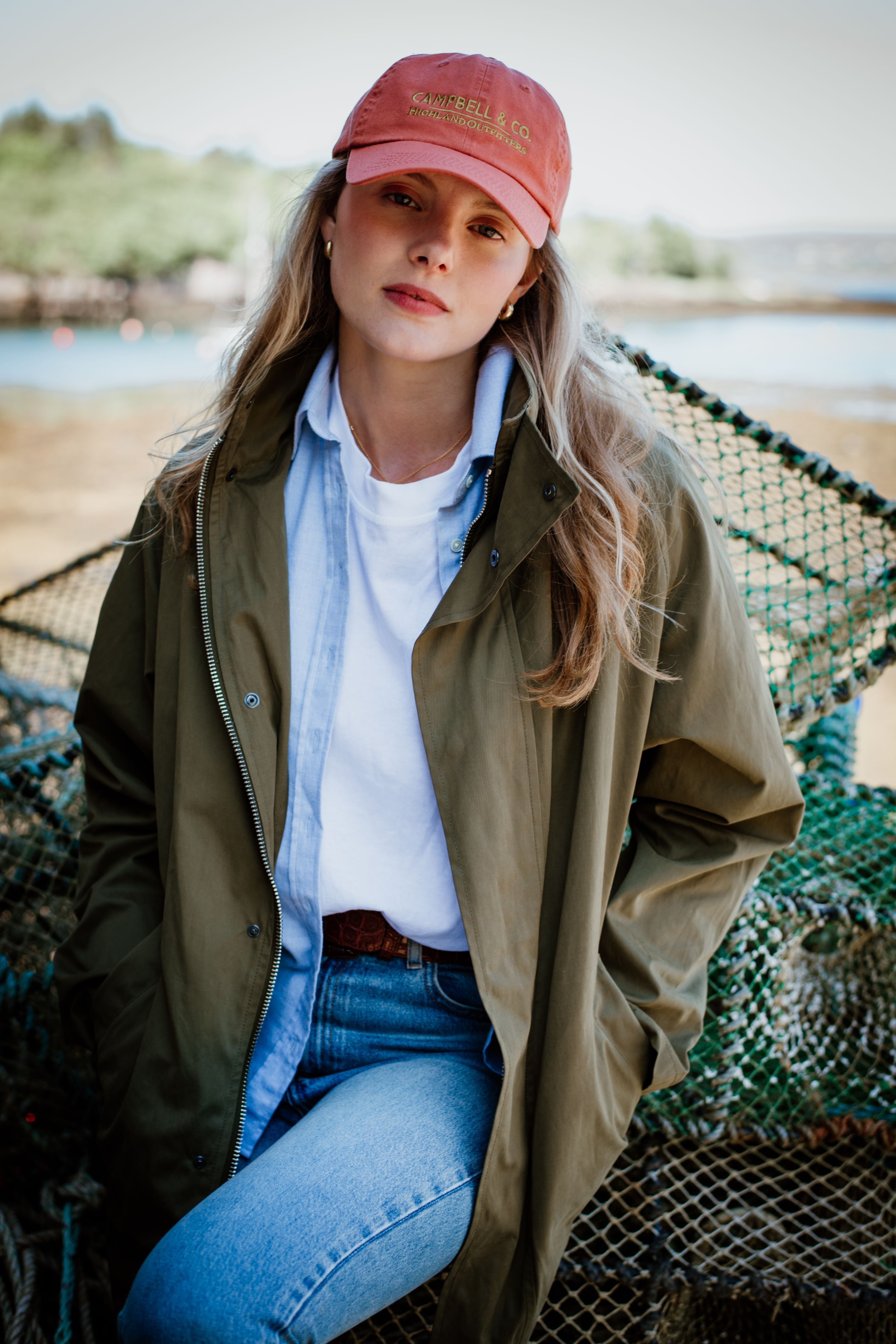 A young woman with long blonde hair wears a red cap, white t-shirt, denim shirt, green jacket, and jeans. She stands outdoors, leaning against stacked fishing nets, with trees and water in the background.