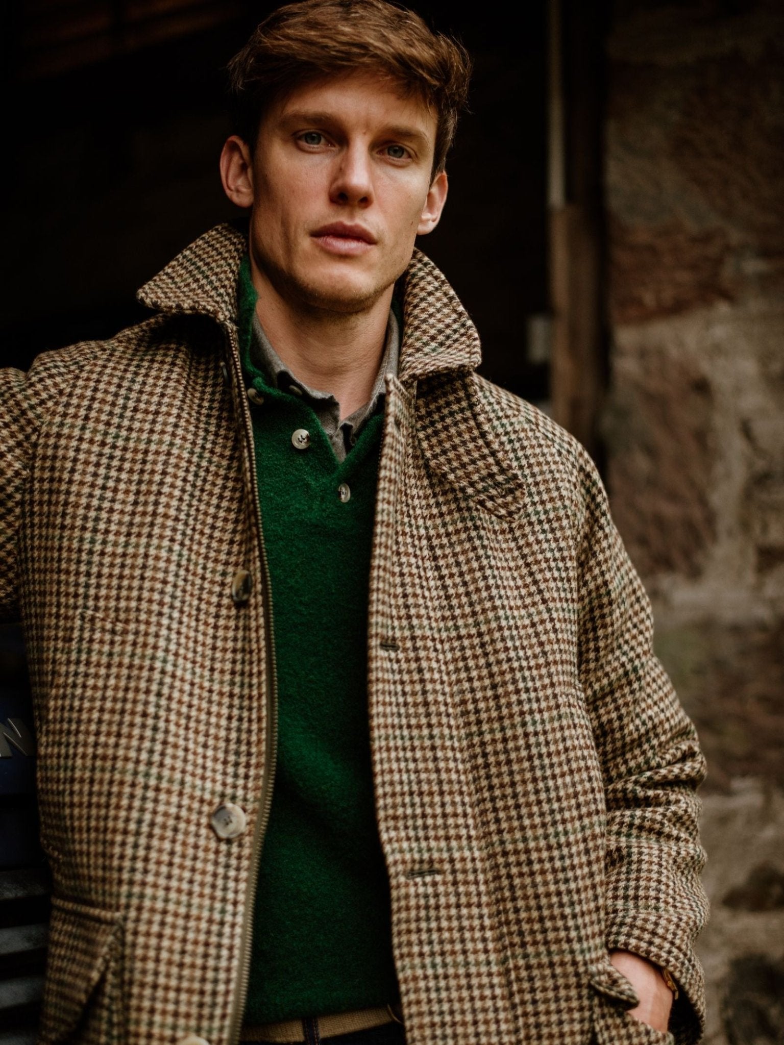 A young man with light brown hair wears the Cannich Coat by Campbell's of Beauly over a green sweater and stands outdoors near a stone wall, looking calmly at the camera.