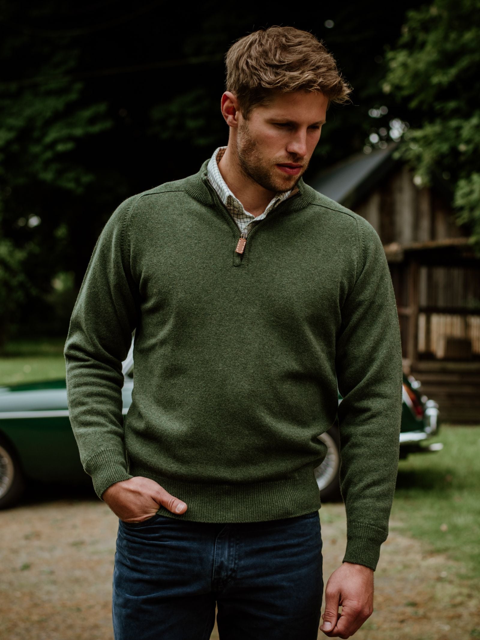 A man with light brown hair and a trimmed beard stands outdoors in the Scottish Borders, wearing Campbells of Beauly Lambswool Quarter Zip Jumper over a plaid shirt and dark jeans, framed by trees and a vintage green car.