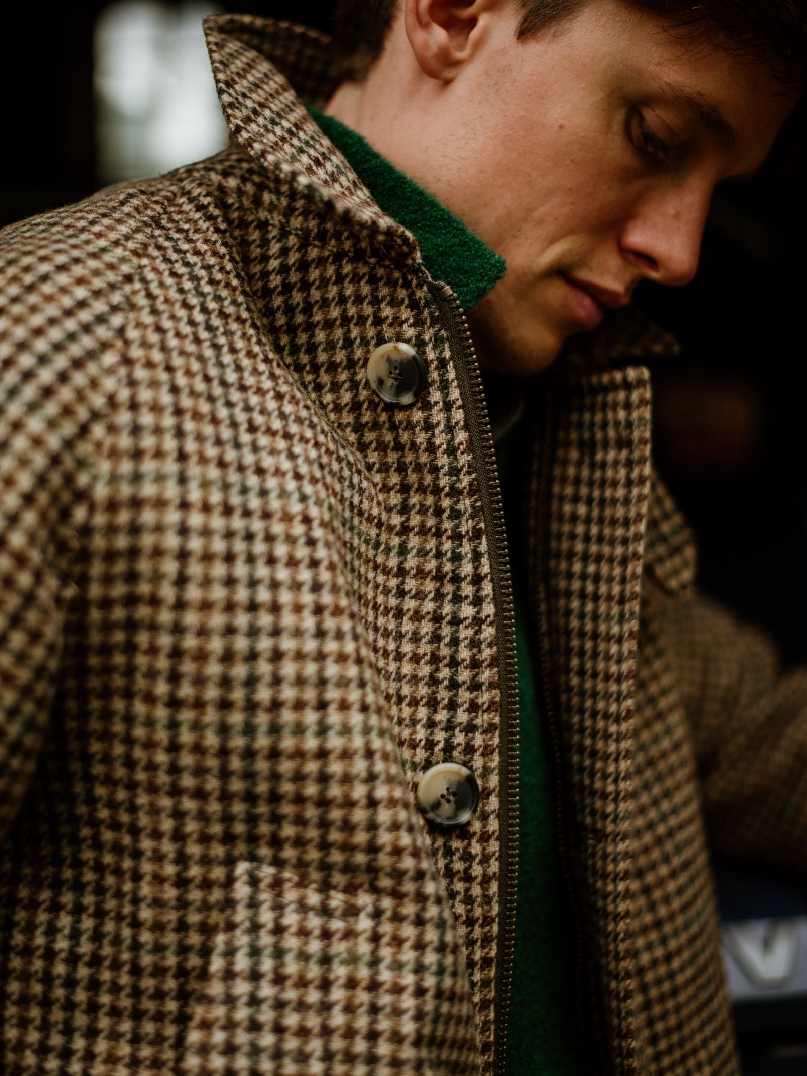A person wears the Campbell's of Beauly Cannich Coat in brown and white houndstooth with large buttons, styled like a classic Loden coat, layered over a green sweater, looking downward in soft lighting.