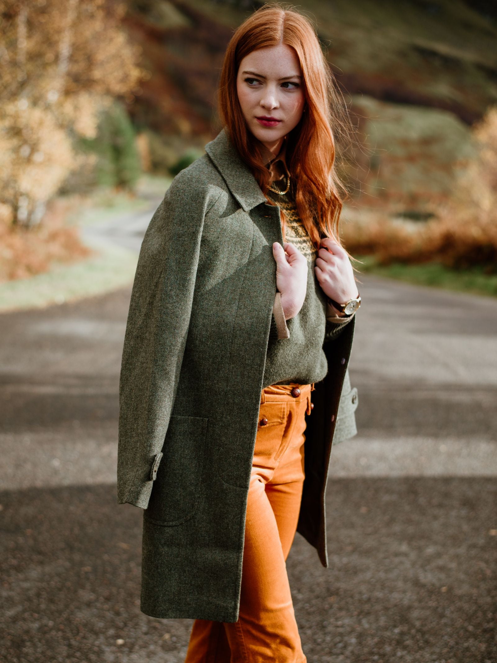 A woman with long red hair wears the Campbells of Beauly Heath Collar Coat and orange pants, standing on a road amid autumn trees and grassy hillsides, highlighting chic everyday style.