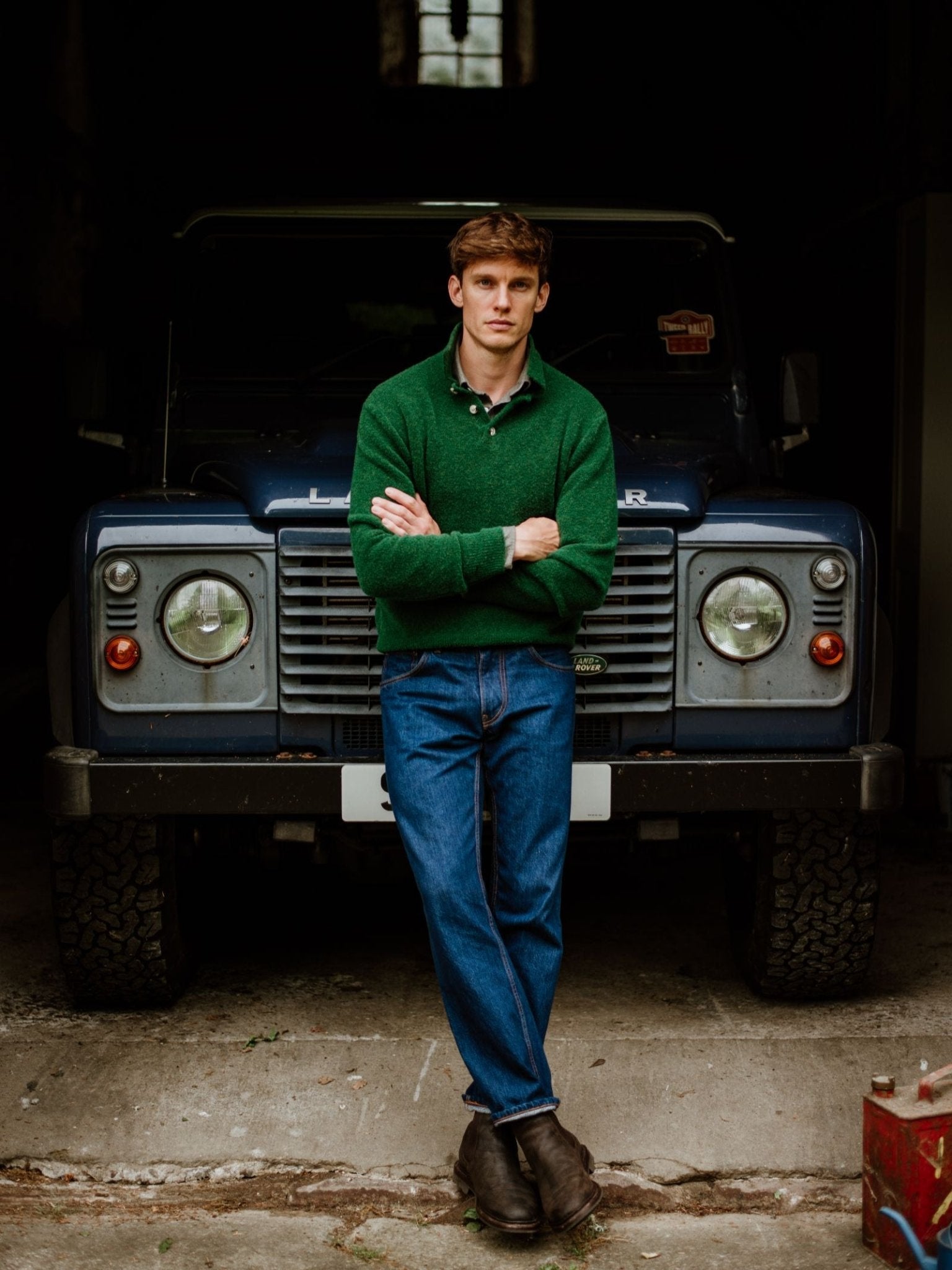 A man wearing a green Campbell's of Beauly Bouclé Polo Sweater and blue jeans stands with arms crossed in front of a dark blue Land Rover inside a dim garage.